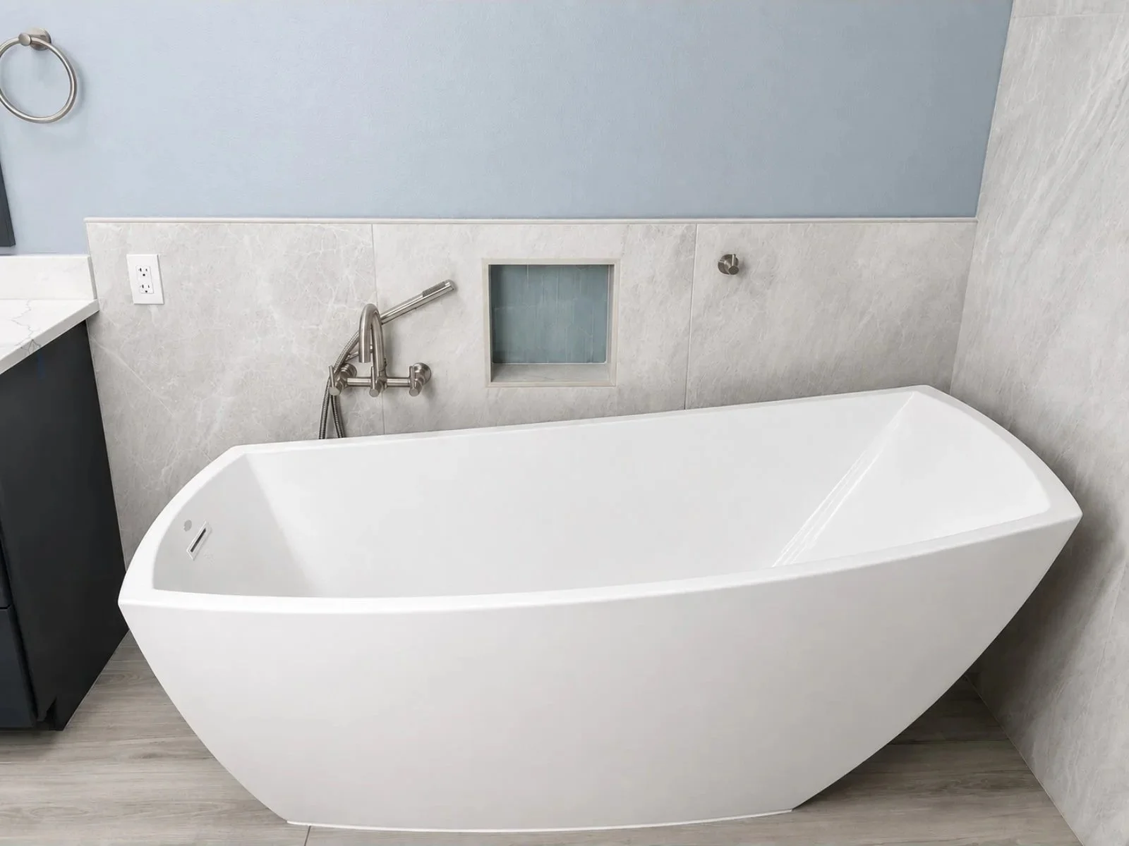 White modern freestanding bathtub in a bathroom with gray wall tiles, a mounted silver faucet with a shower hose, a small square recessed shelf, and a gray wood-like floor.