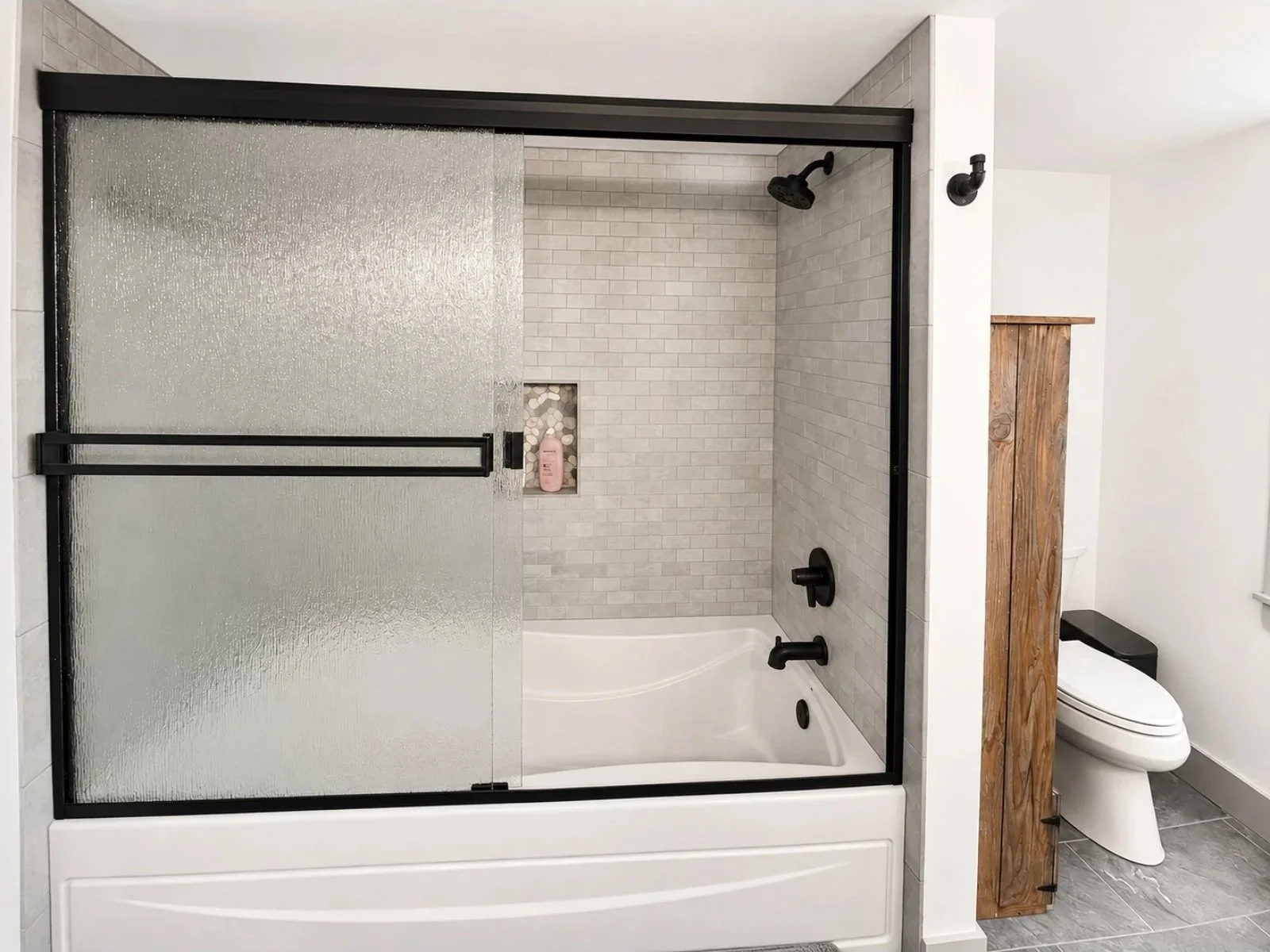 Bathroom with a shower and a toilet, featuring a frosted glass sliding door, gray tile walls, black fixtures, and a wood partition.