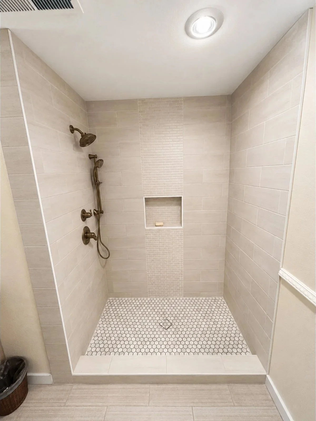 A modern walk-in shower with beige tile walls, a niche for toiletries, a ceiling light, and a dual shower head with a handheld option.
