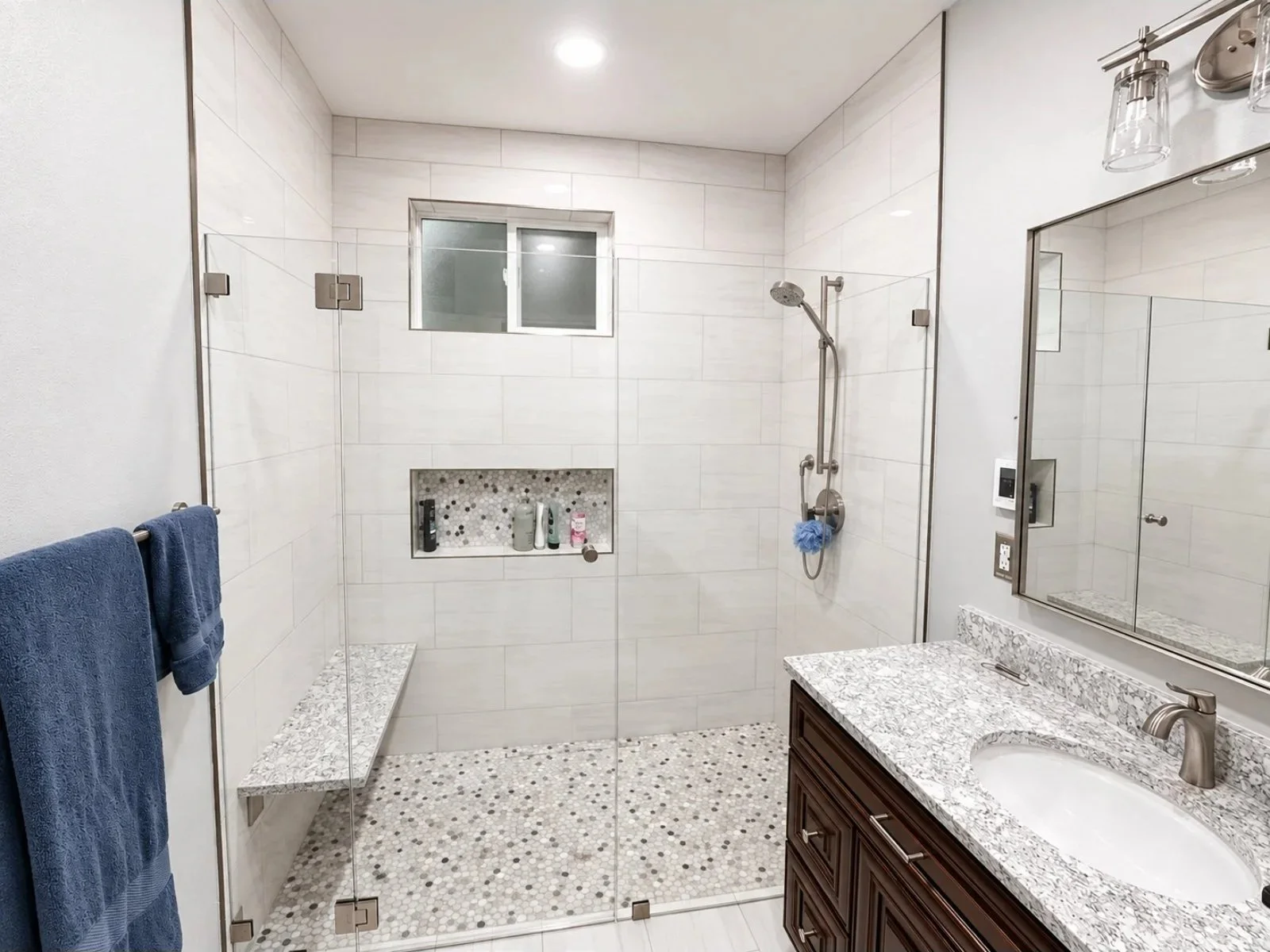 Modern bathroom with glass shower enclosure, white tiled walls, small mosaic tile floor, medicine cabinet with mirror, granite countertop, dark wooden vanity, and a window.