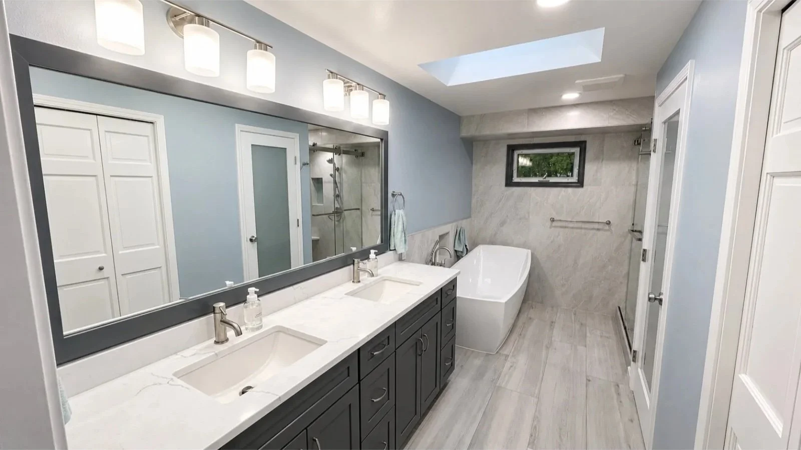 Modern bathroom with dual sinks, large mirror, bathtub, shower area, skylight, and light blue walls.
