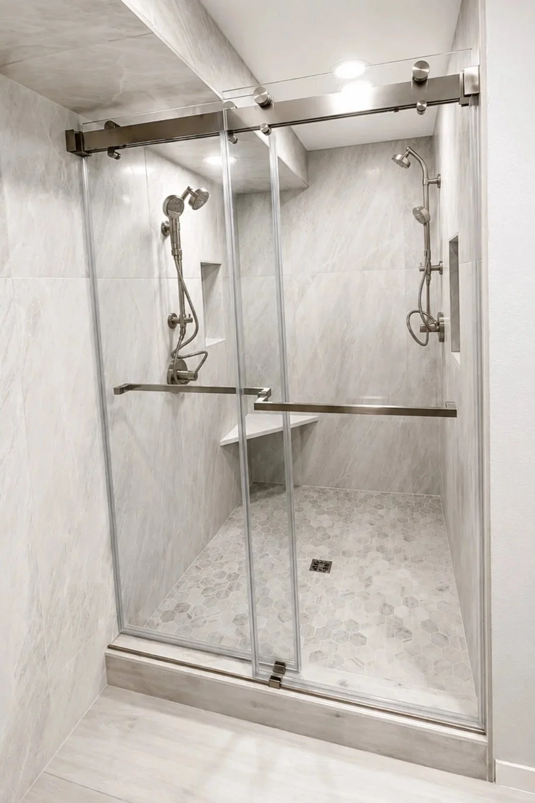 A modern glass-enclosed shower with two handheld shower heads, built-in wall niche, and a bench in a minimalist bathroom with white marble walls and hexagonal tile floor.
