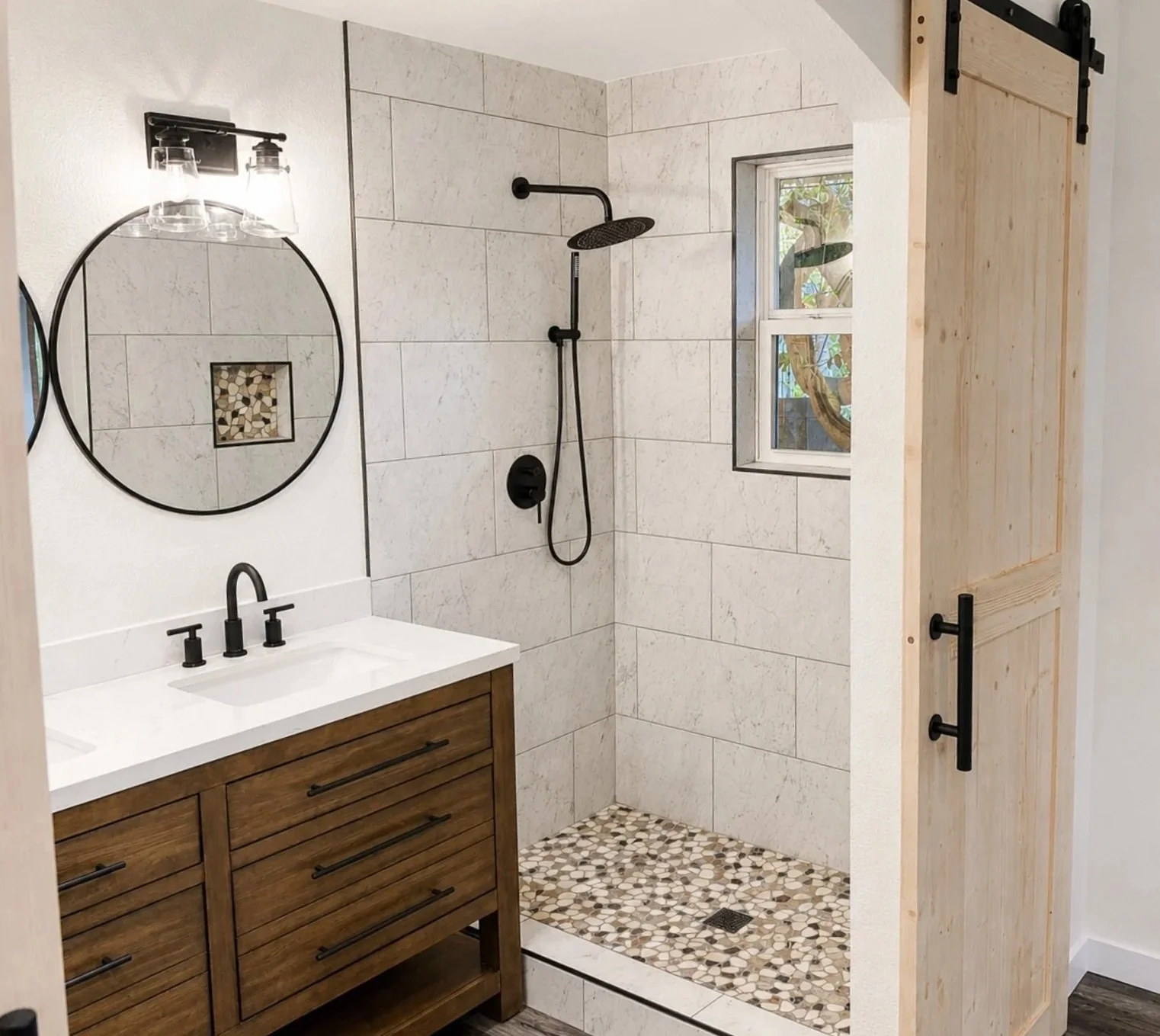 Bathroom with walk-in shower featuring black fixtures, a modern wooden vanity with a white countertop, two round mirrors, and a rustic sliding barn door.