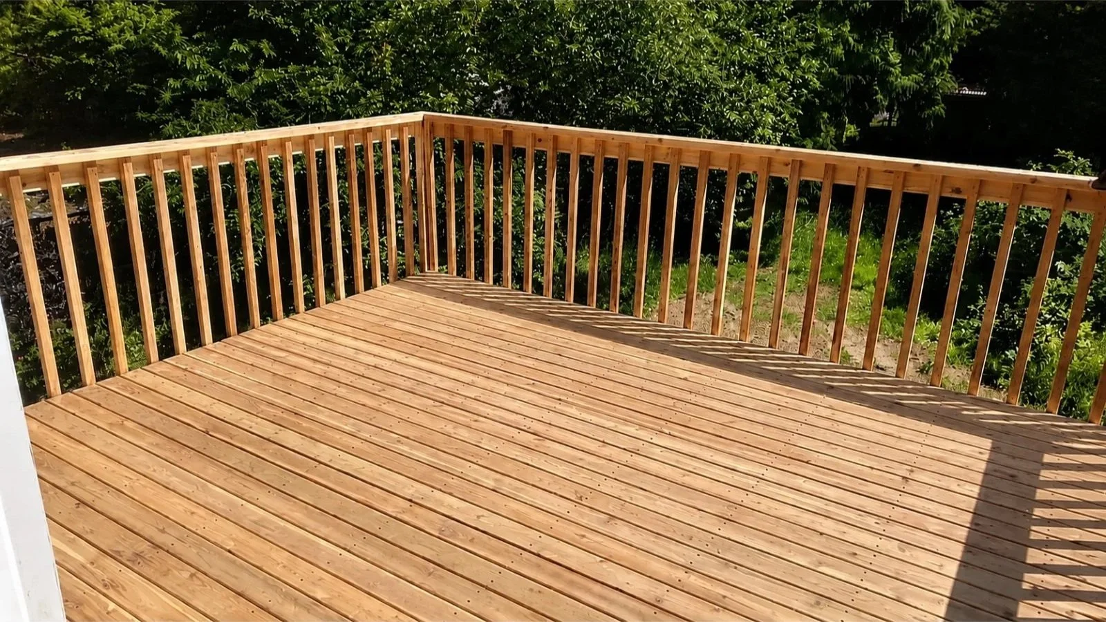 Newly built wooden deck with railing, overlooking a green backyard with bushes and trees.