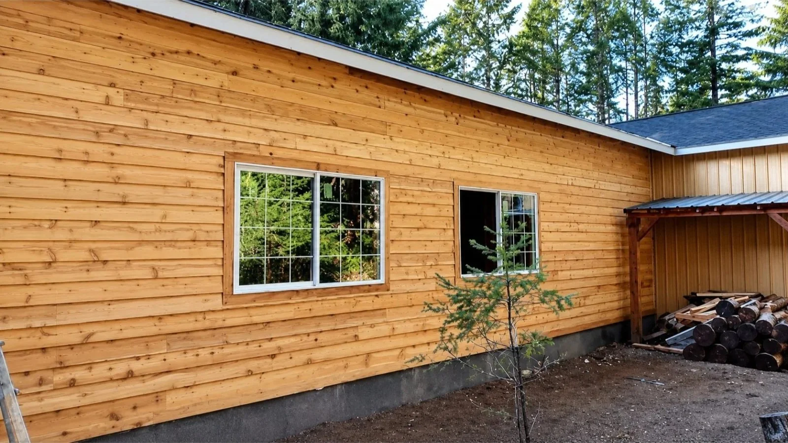 A newly built wooden house with horizontal wooden siding, two windows with grid panes, and a small tree in front. There is a stack of logs under a metal-roofed shelter on the right side.