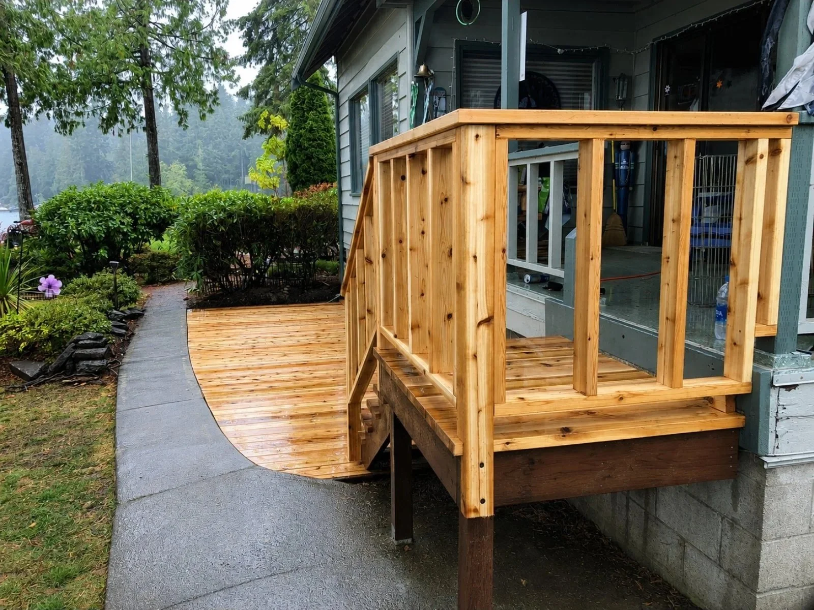 Newly built wooden deck with railing attached to house, with a concrete walkway and garden with flowering plants nearby.
