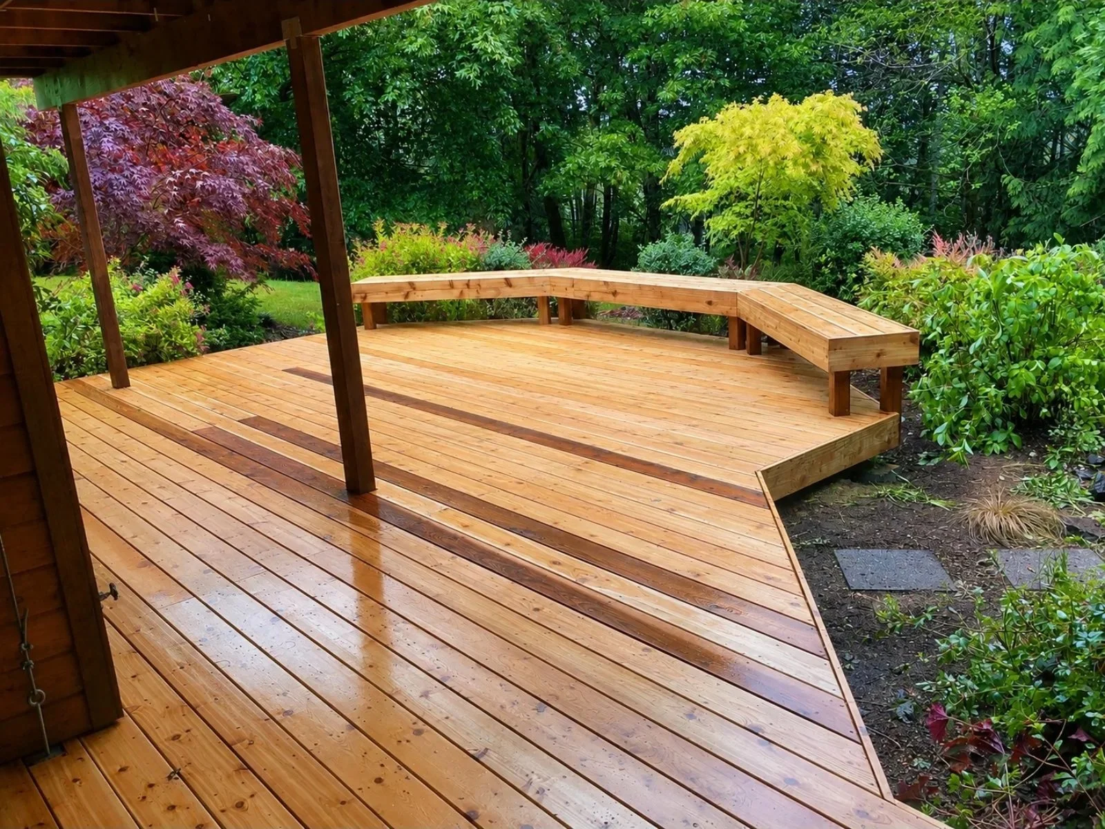 A newly built wooden deck with a built-in bench and a garden surrounding it, featuring colorful trees and shrubs.