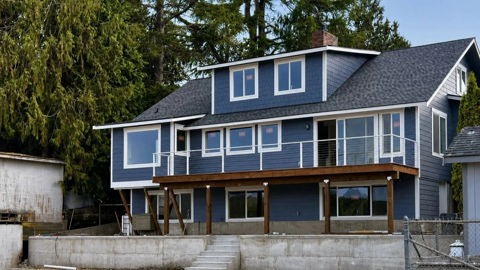A two-story house with blue siding and large windows. The house has a deck on the second floor with white railing and a staircase leading down to the ground level. There are trees in the background.