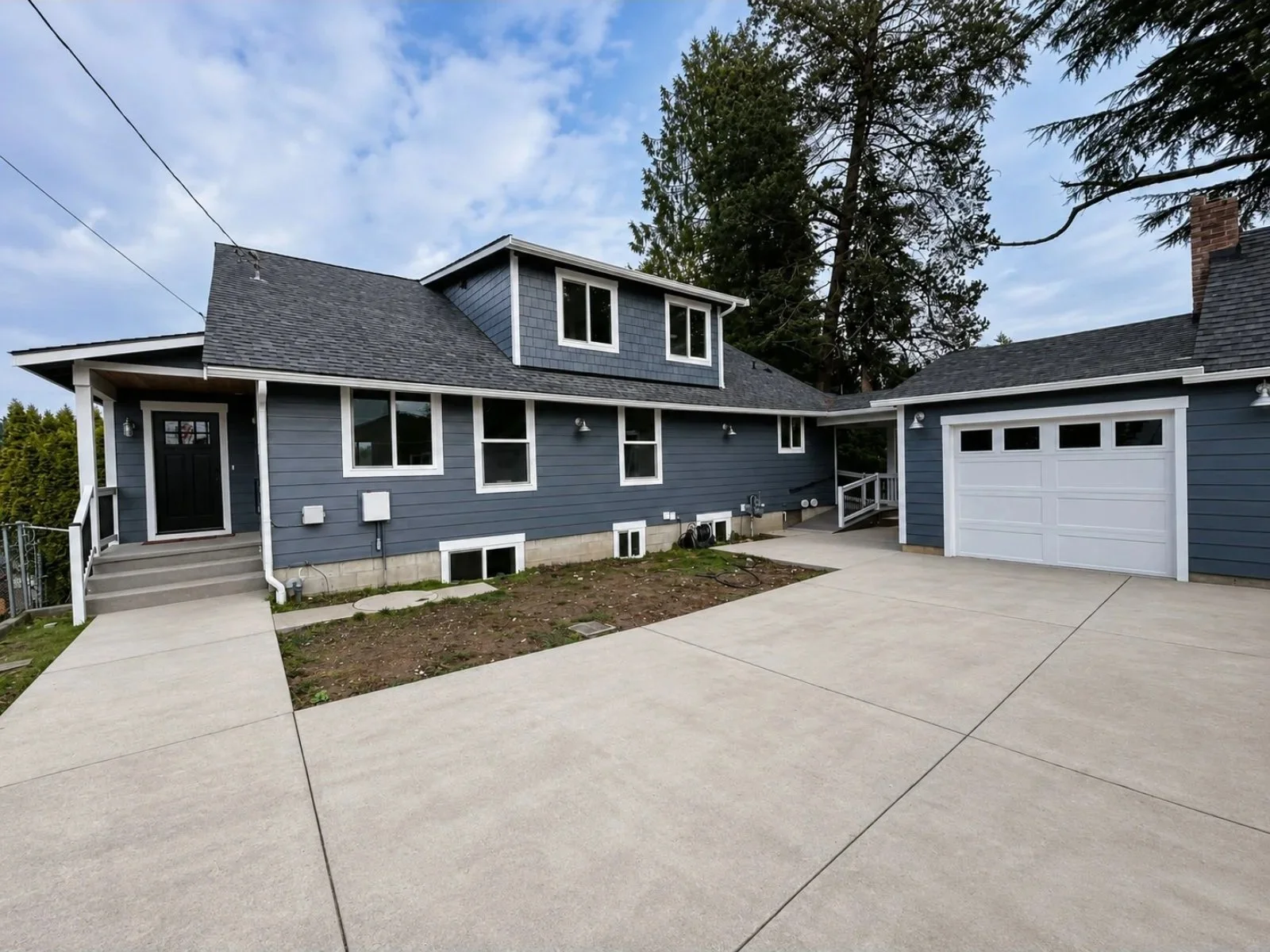 Front view of a newly sided and painted blue house with a driveway leading to a white garage door, with a small porch and steps at the entrance, surrounded by trees and a partly cloudy sky. Brand new windows visible. 