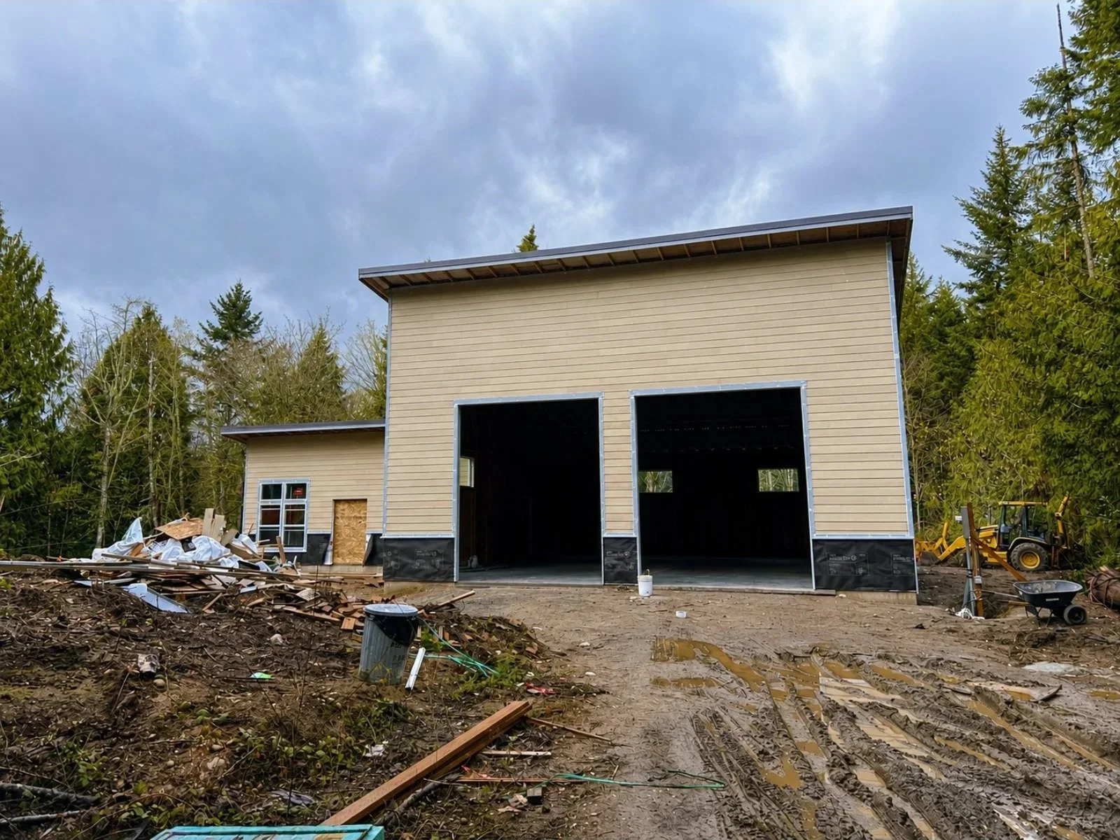 A house under construction with beige siding, two large open garages, and a small window on the left side. The surrounding area has construction debris, piles of dirt, and construction equipment, with a backdrop of trees and a cloudy sky.