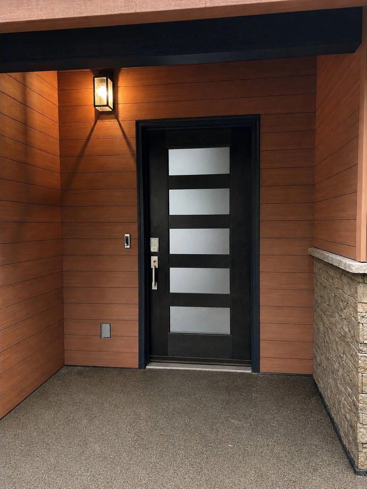 Modern front door with black frame and frosted glass panels, wood-paneled wall, stone accent wall, wall-mounted light fixture, and textured carpet.