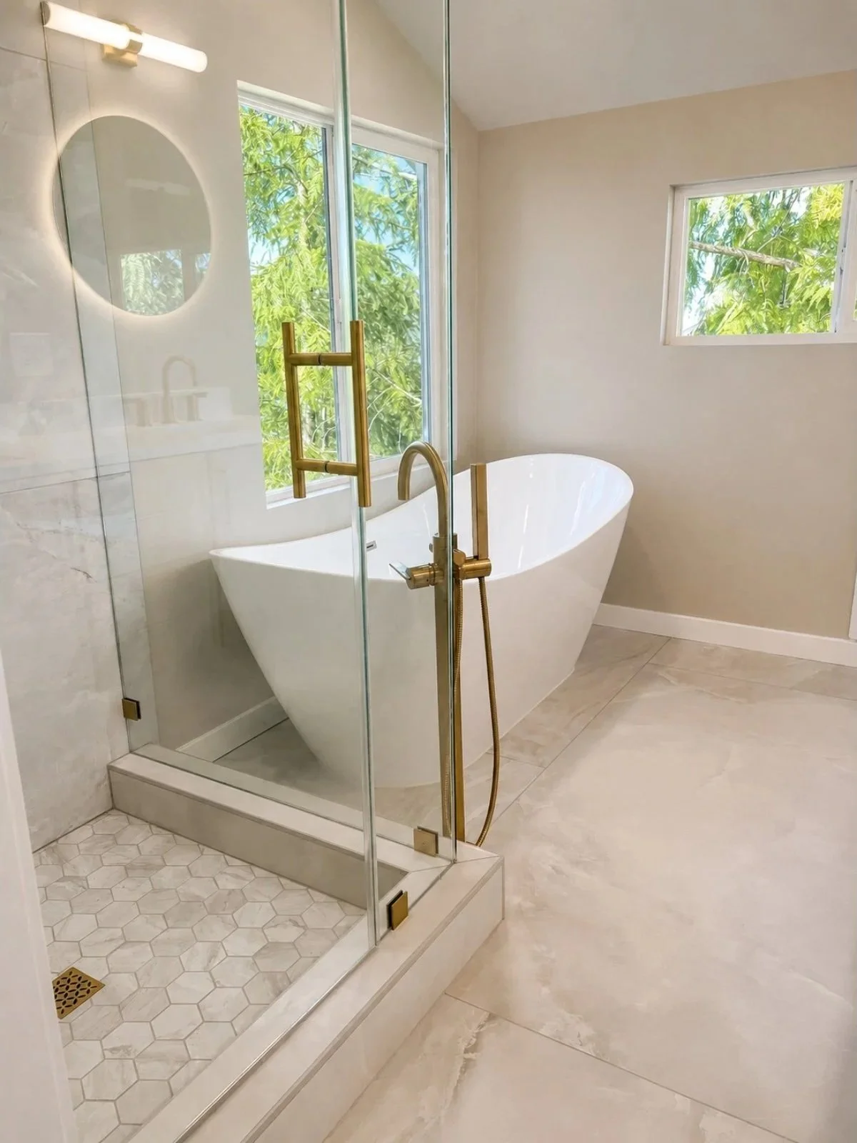 Modern bathroom with freestanding bathtub, glass shower enclosure with brass fixtures, large windows revealing green trees, and beige marble tile flooring.