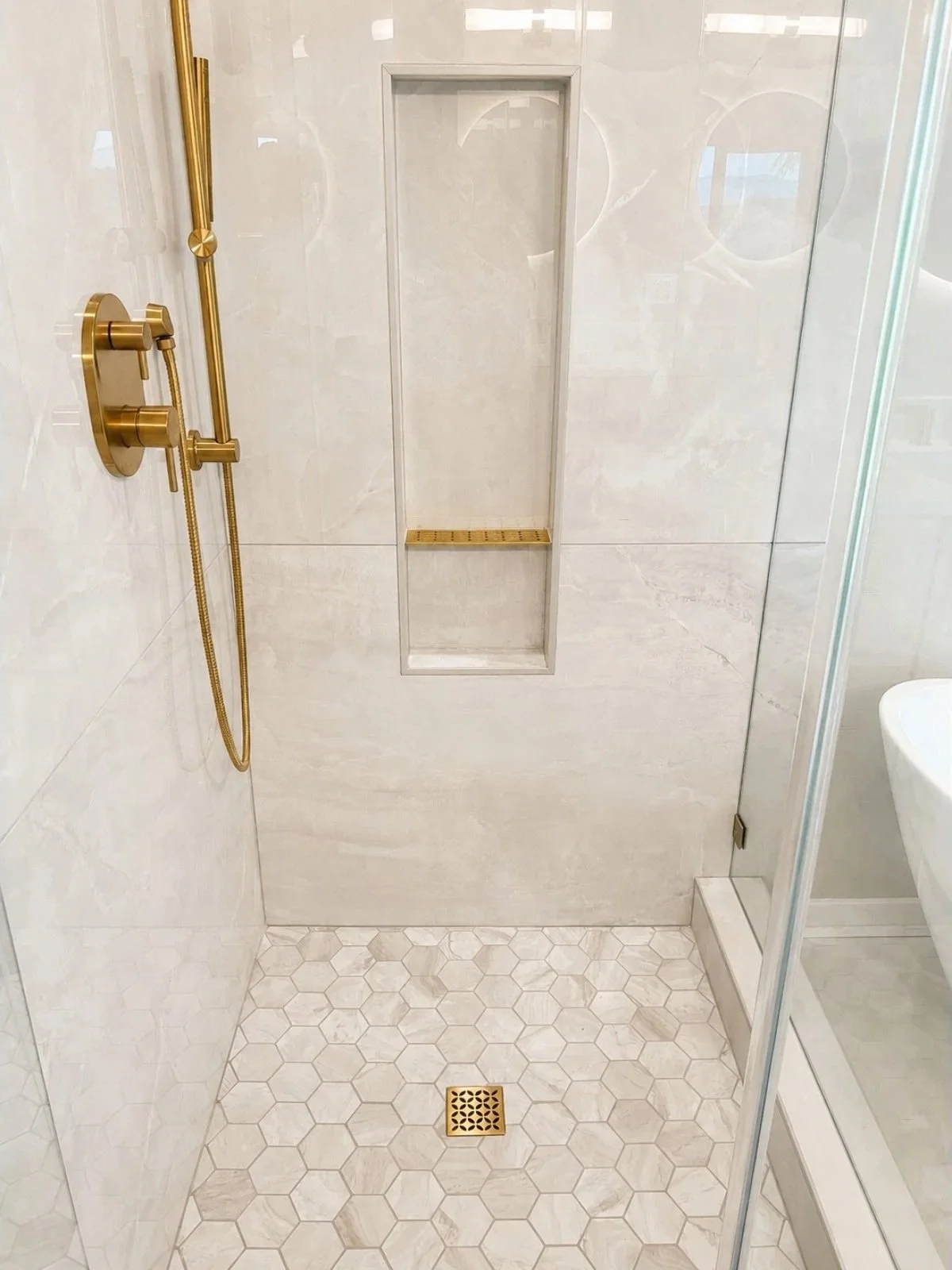 A modern walk-in shower with a gold shower head and controls on the left wall, a built-in niche with a wooden shelf for toiletries, a floor drain with a decorative cover, and a glass shower door on the right. The walls and floor are made of cream-col