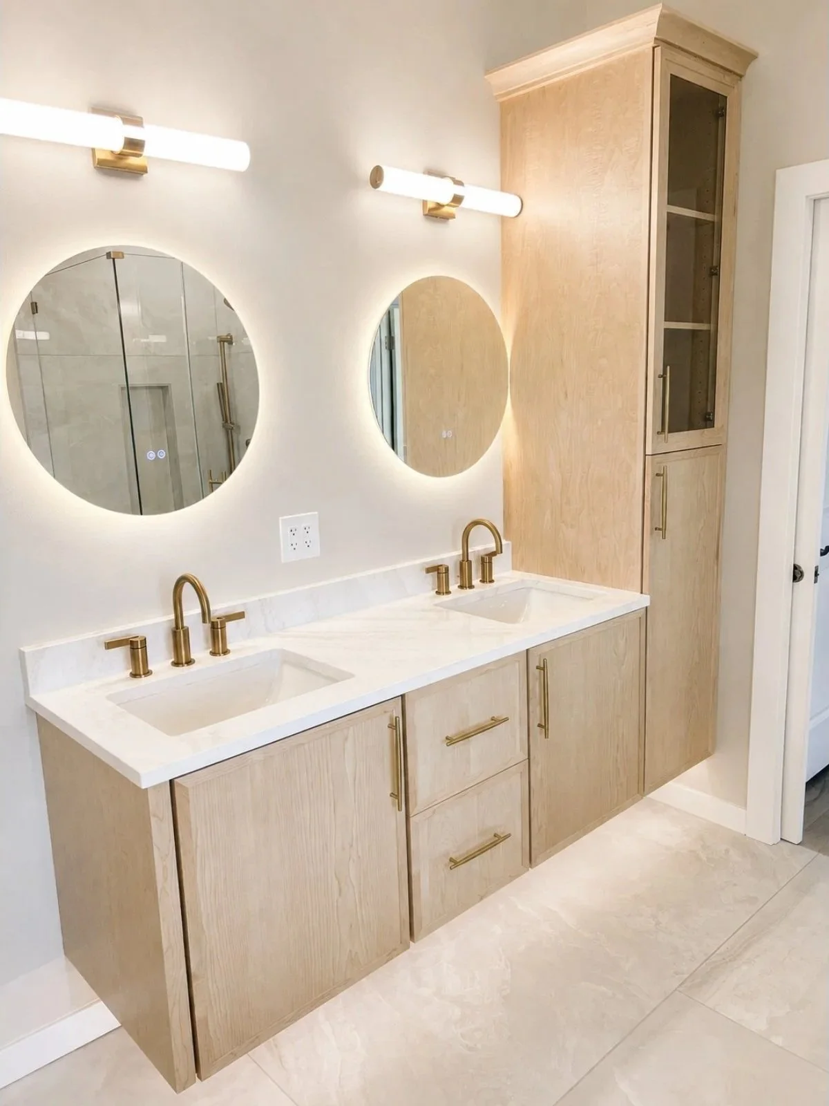 Modern bathroom with dual sinks, light wood cabinets, and round mirrors. Gold fixtures and a tall storage cabinet are visible.