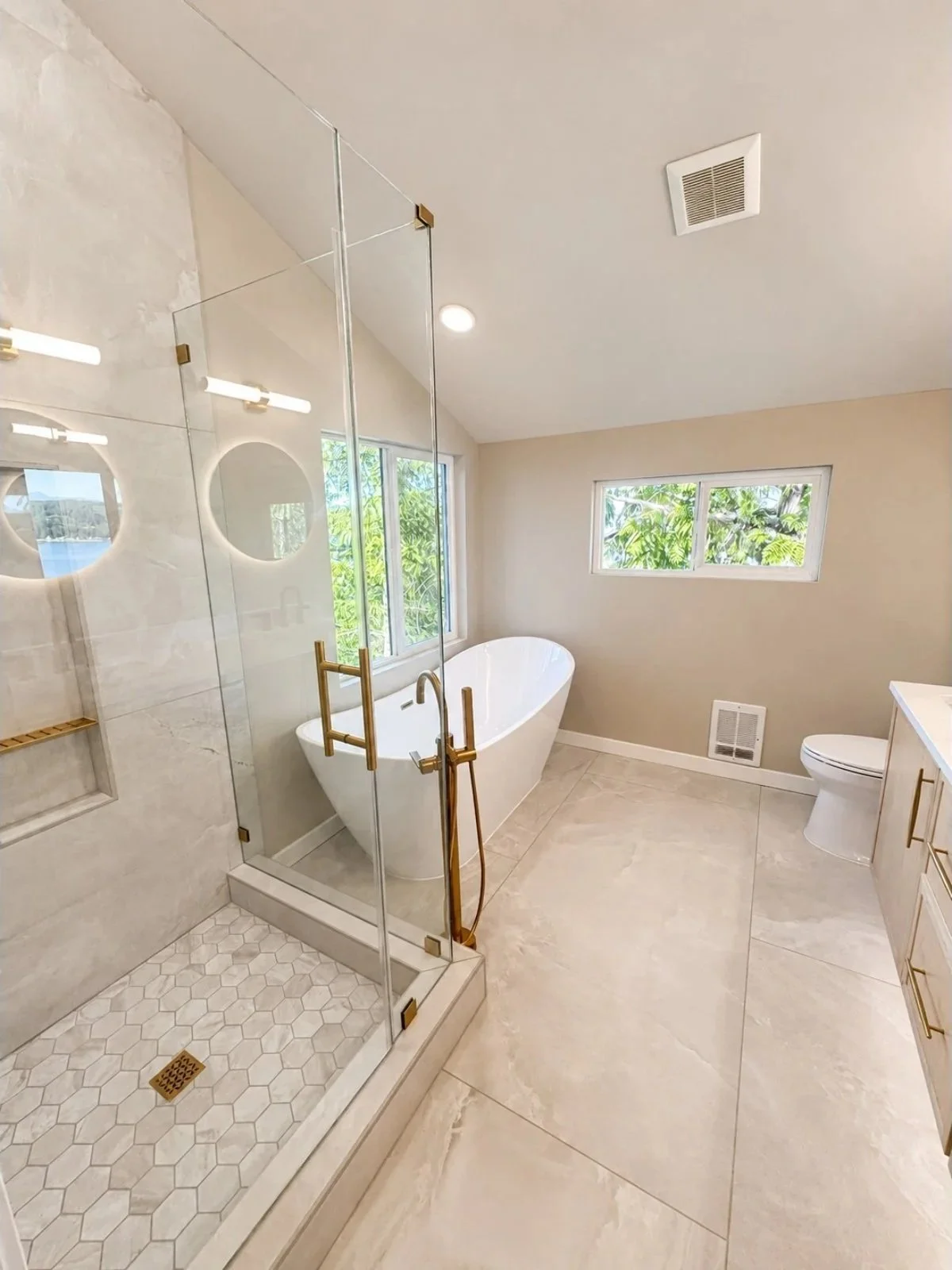 Modern bathroom featuring a glass-enclosed shower with brass fixtures, a white freestanding bathtub, a window with greenery outside, a beige toilet, and a stone tile floor.