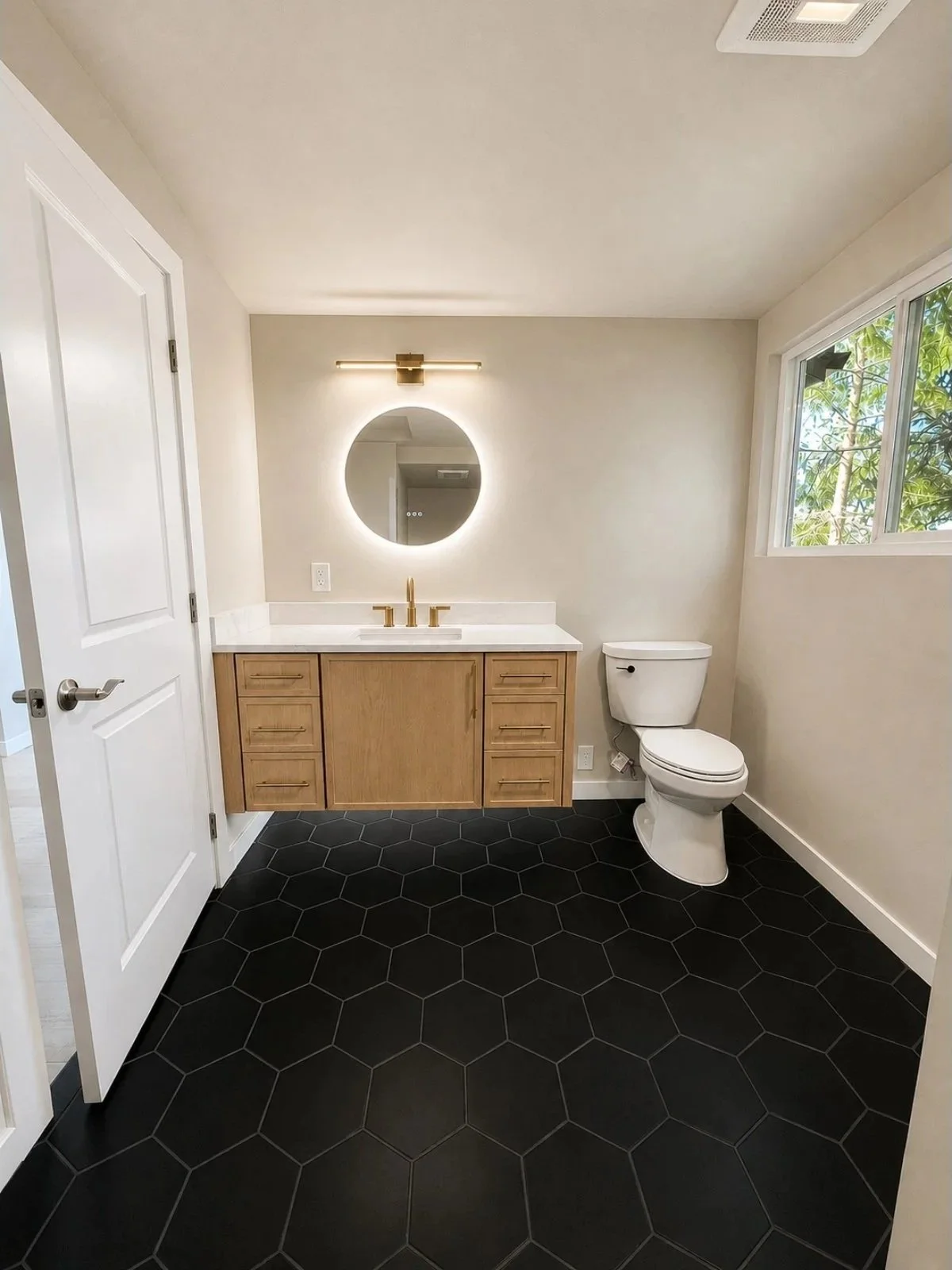 Modern bathroom with black hexagonal floor tiles, wooden vanity with gold fixtures, round backlit mirror, and a white toilet beside a window with greenery outside.
