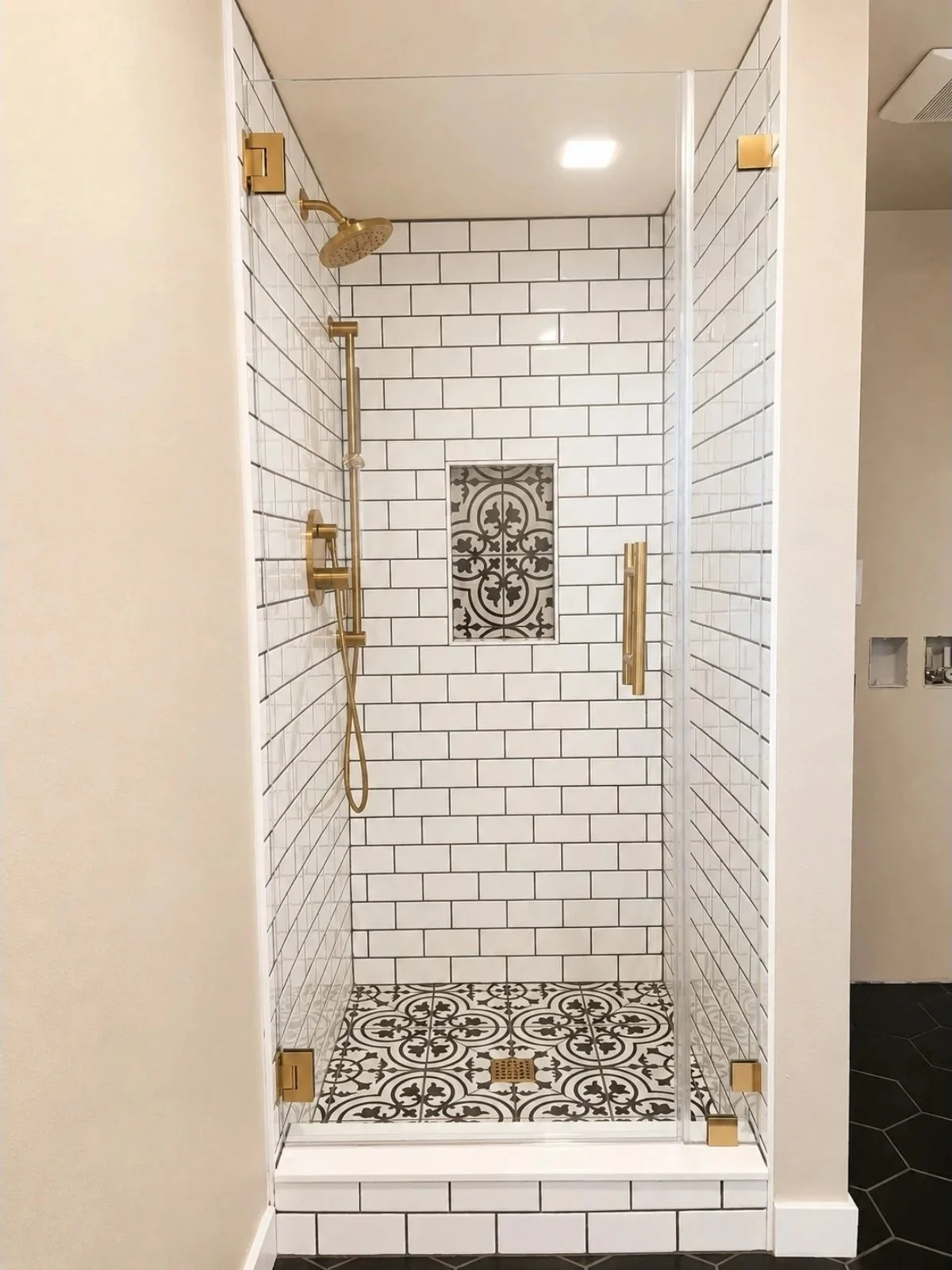 A modern walk-in shower with white subway tiles, black grout, and ornate black and white patterned floor tiles. It has gold fixtures, including a rain showerhead, handheld shower, and a vertical grab bar. There's a niche with decorative black and whi
