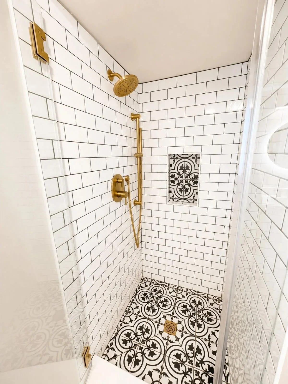 Shower with white subway tile walls, black and white patterned floor tiles, and brass fixtures including a showerhead, handle, and grab bar.