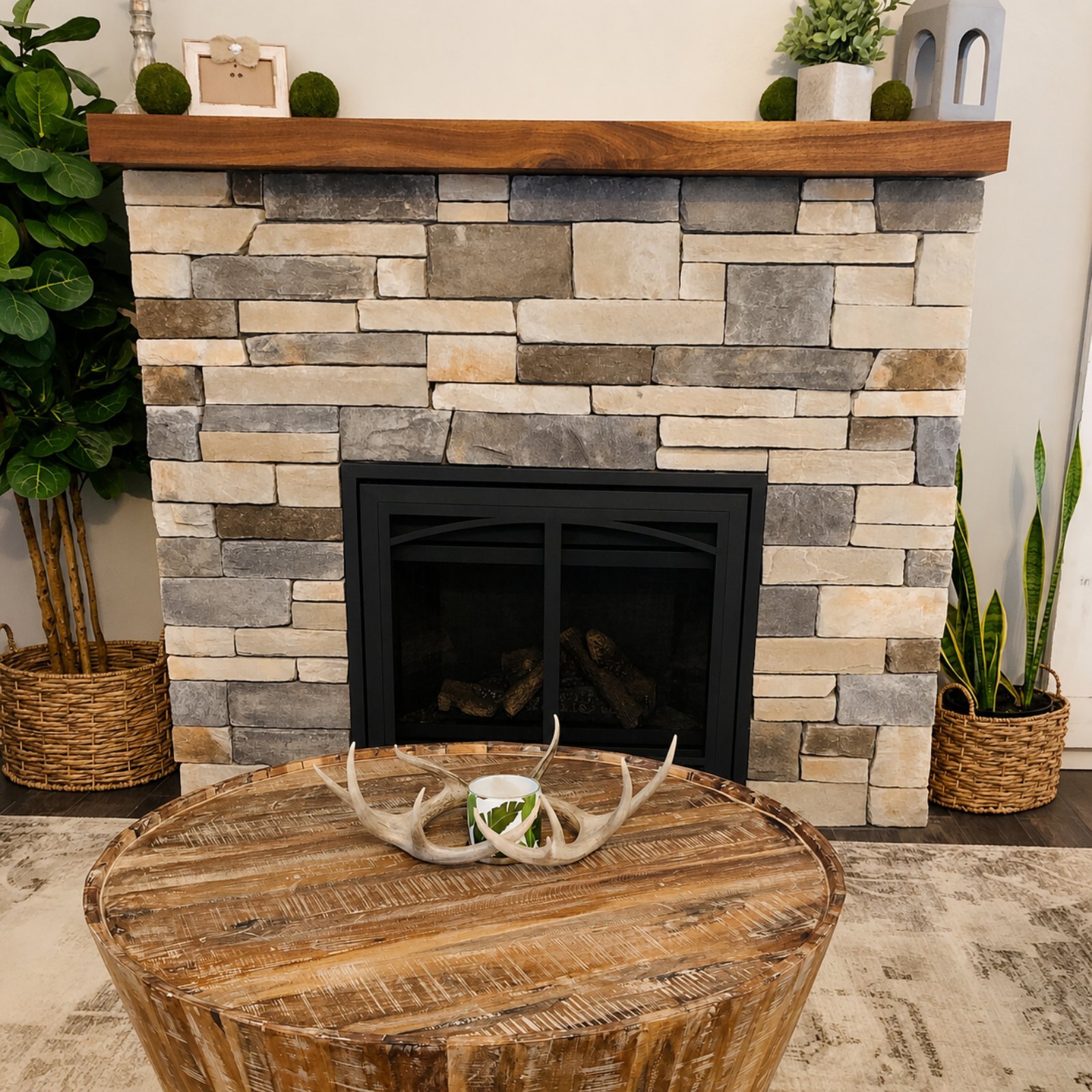 A cozy living room fireplace with a stacked stone facade, decorated with greenery and white ornaments on top. A large wooden mantel sits above the fireplace. In front, a round wooden table holds a decorative antler centerpiece with a green plant and 