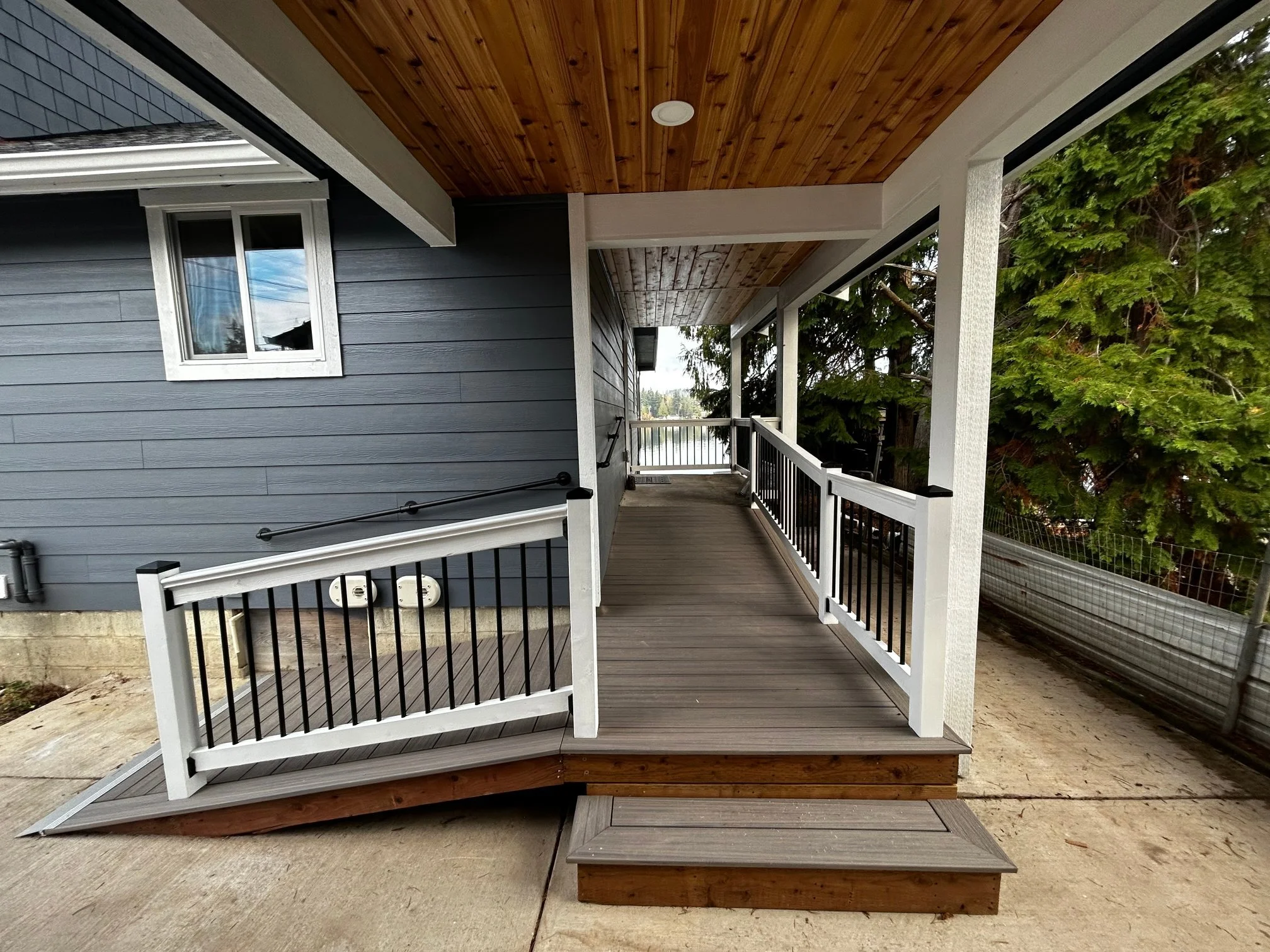 A house porch with wooden flooring, a wooden ceiling, and white trim. There is a railing with white posts and black balusters, and stairs leading down. A window with white trim is on the house's dark blue siding, and trees are visible in the background.