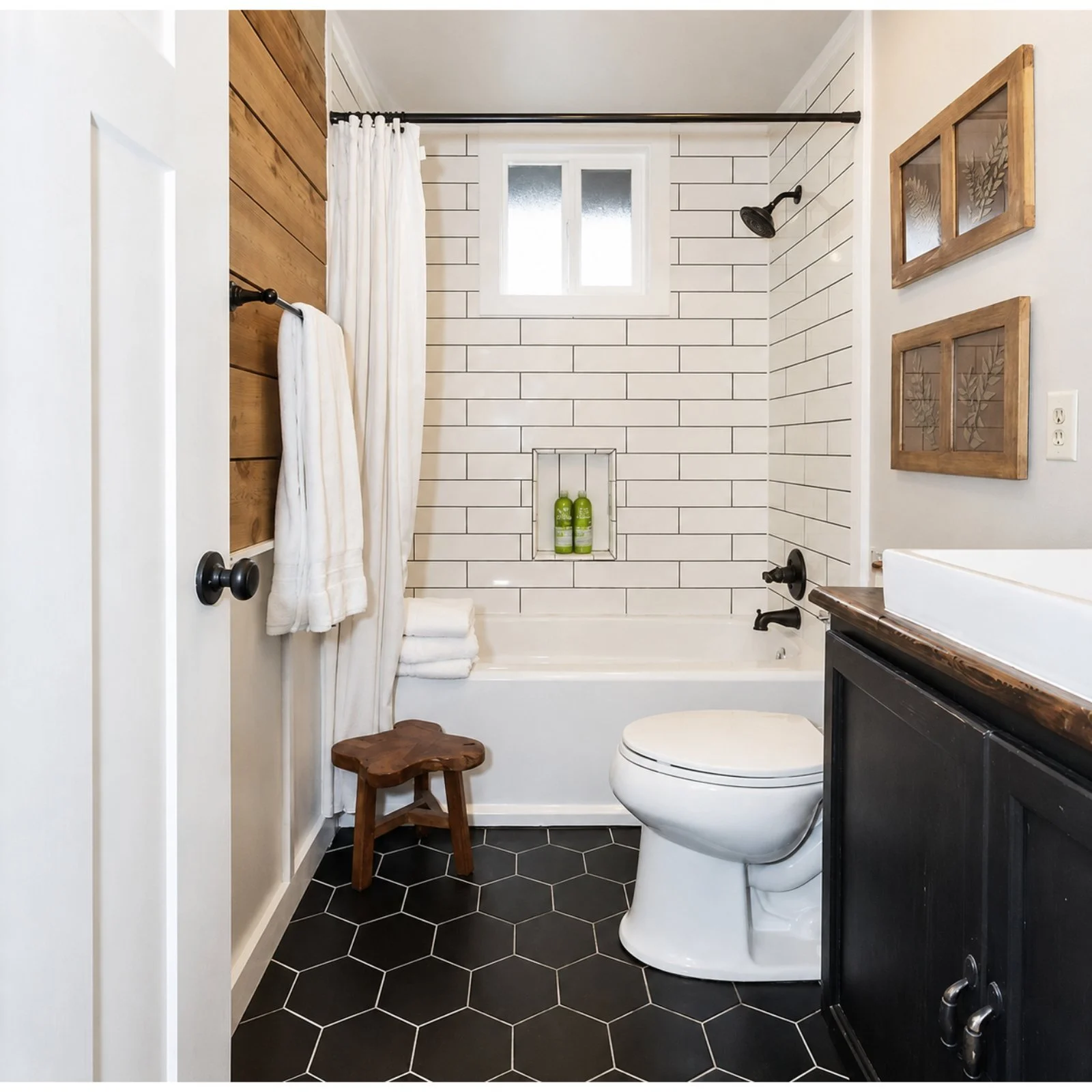 Modern bathroom with black hexagon tile floor, white brick walls, small window, wood accents, and a walk-in shower with white curtain and built-in niche with green bottles.
