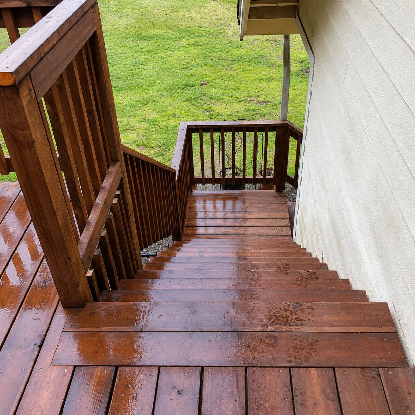 Wooden staircase outside from upper level looking downward, with a handrail on the left, a white wall on the right, and a grassy yard with patches of dirt visible at the bottom.