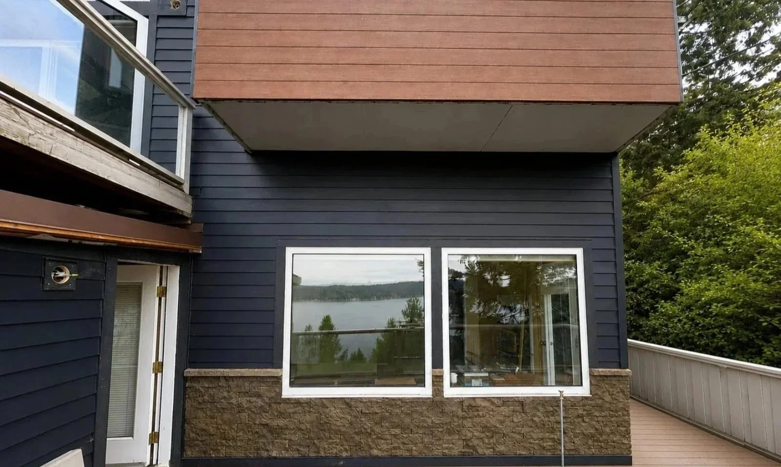 Exterior of a modern house with dark blue siding, large windows, and a balcony with a view of a lake and trees in the background.