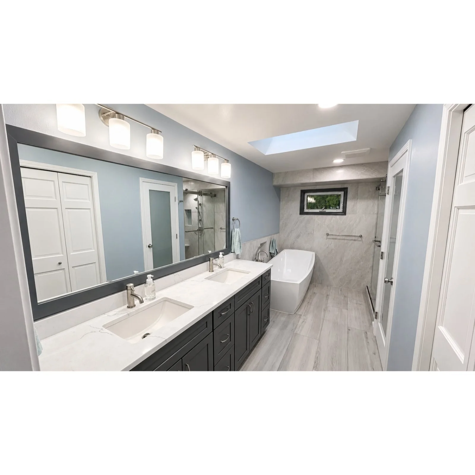 Modern bathroom with double sink vanity, large mirror, wall-mounted lights, freestanding bathtub, walk-in shower, small window, and skylight, with gray and white color scheme.