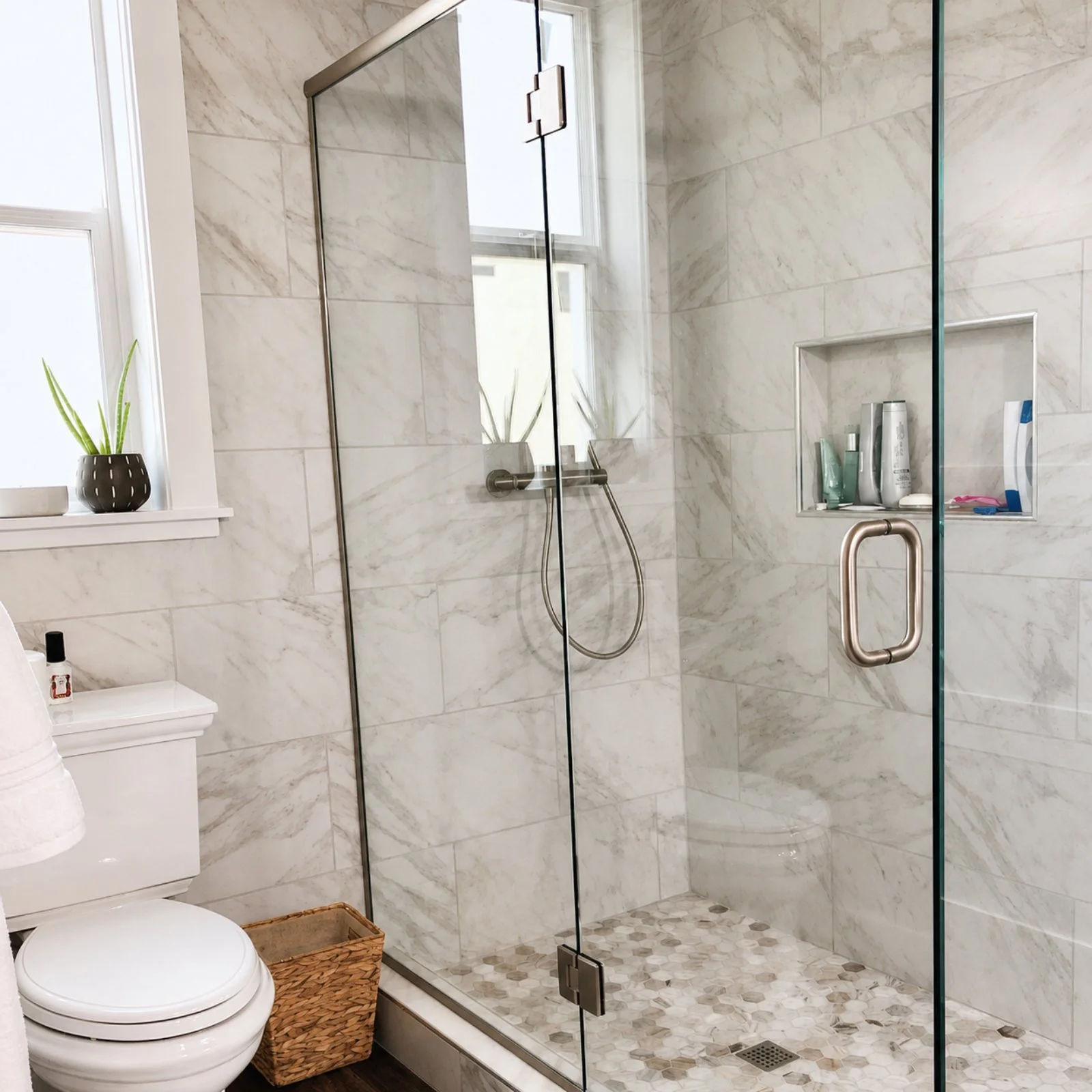 A modern bathroom featuring a walk-in shower with glass door and marble tiled walls, built-in shower niche with toiletries, a small window with a potted plant, and a toilet with a wicker basket nearby.