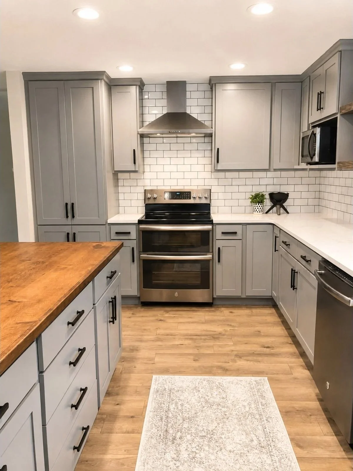 Modern kitchen with gray cabinets, white subway tile backsplash, stainless steel oven and range hood, wooden floor, and a wooden countertop island. Small potted plant and black decorative items on the countertop.