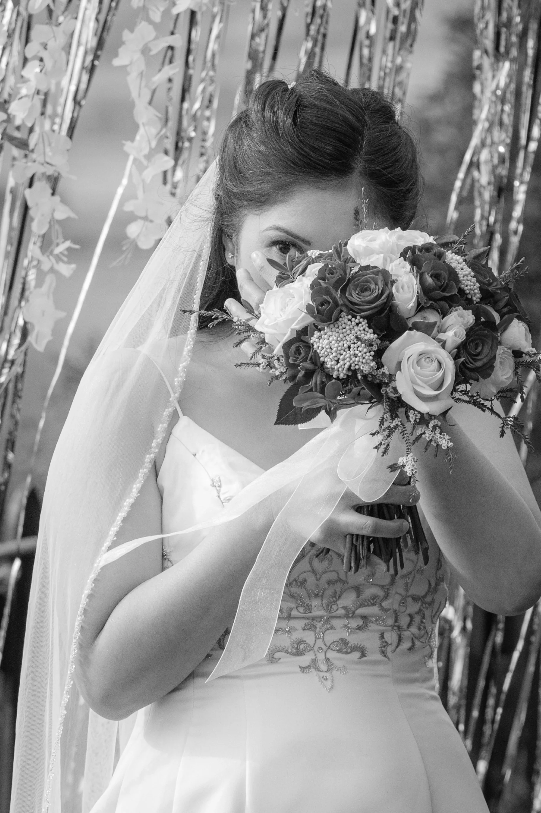 Black and white photo of a bride holding a bouquet of flowers, covering part of her face, with decorative background.