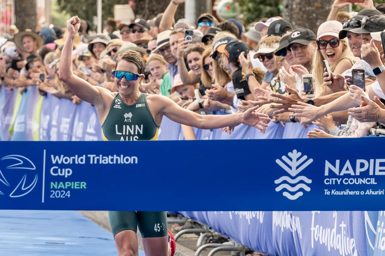 Female triathlon athlete crossing the finish line, smiling with her right arm raised, crowd cheering behind a blue banner at the 2024 World Triathlon Cup in Napier, New Zealand.