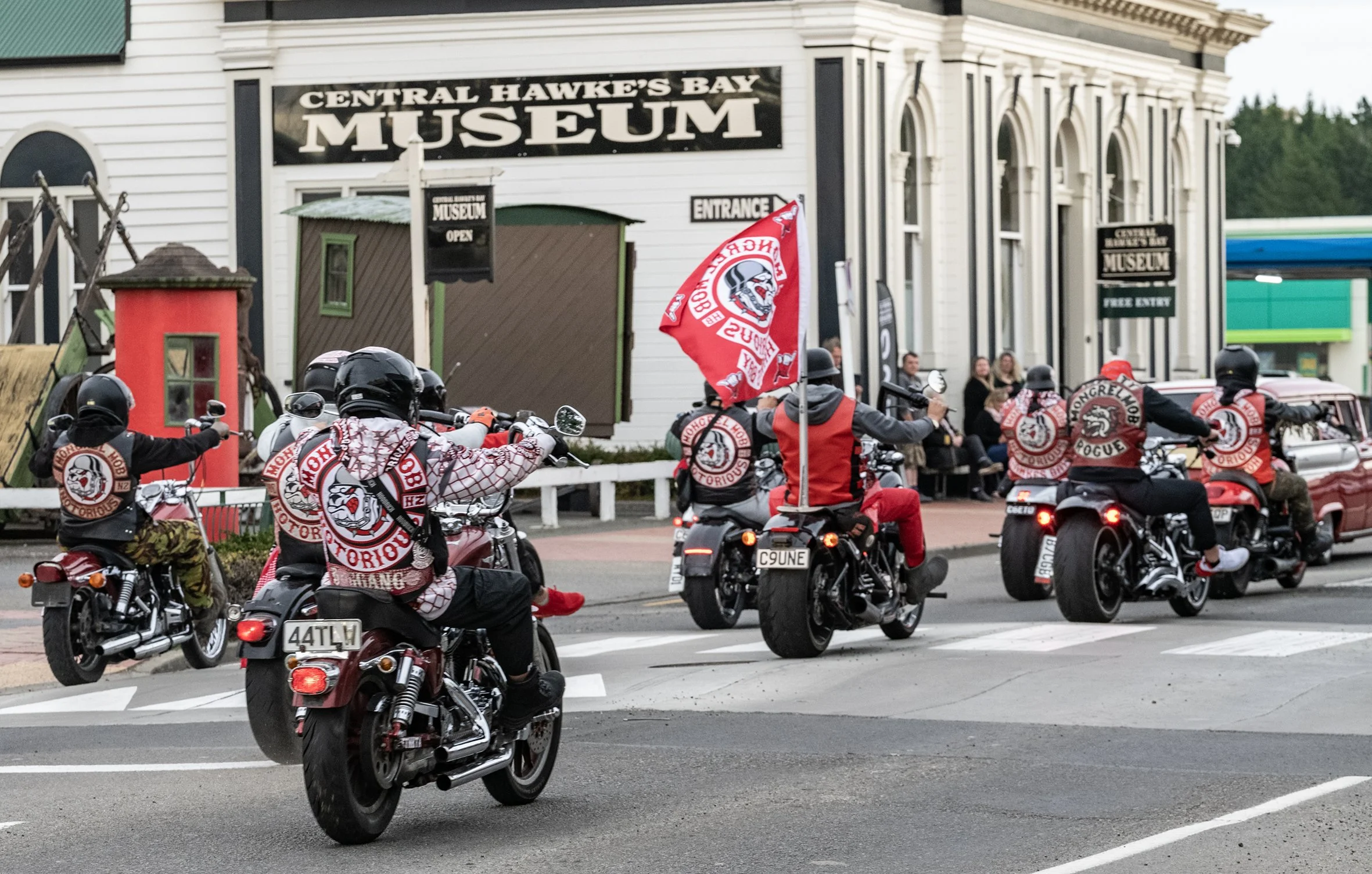 A group of motorcyclists riding past the Central Hawke's Bay Museum, with some wearing red and black jackets and helmets, and holding flags.