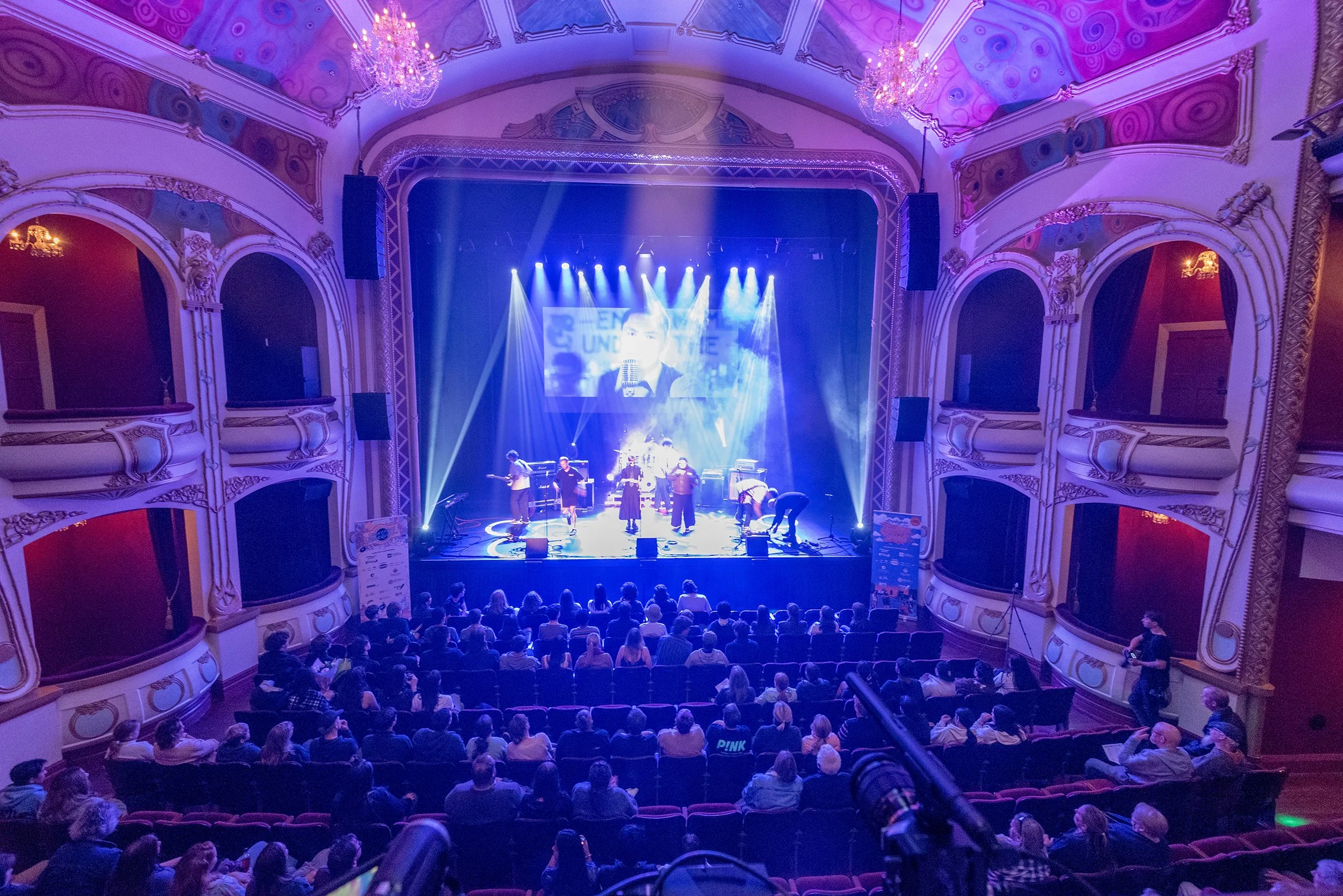 A theater with an ornate decorated interior and chandeliers, showing a live music performance on stage with multiple musicians and a large screen in the background.