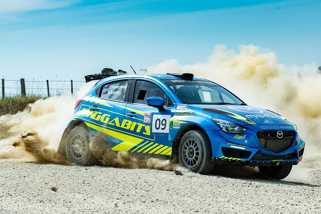 A blue Mazda rally car with yellow graphics and sponsor logos, number 09, kicking up dust on a gravel road during a race.