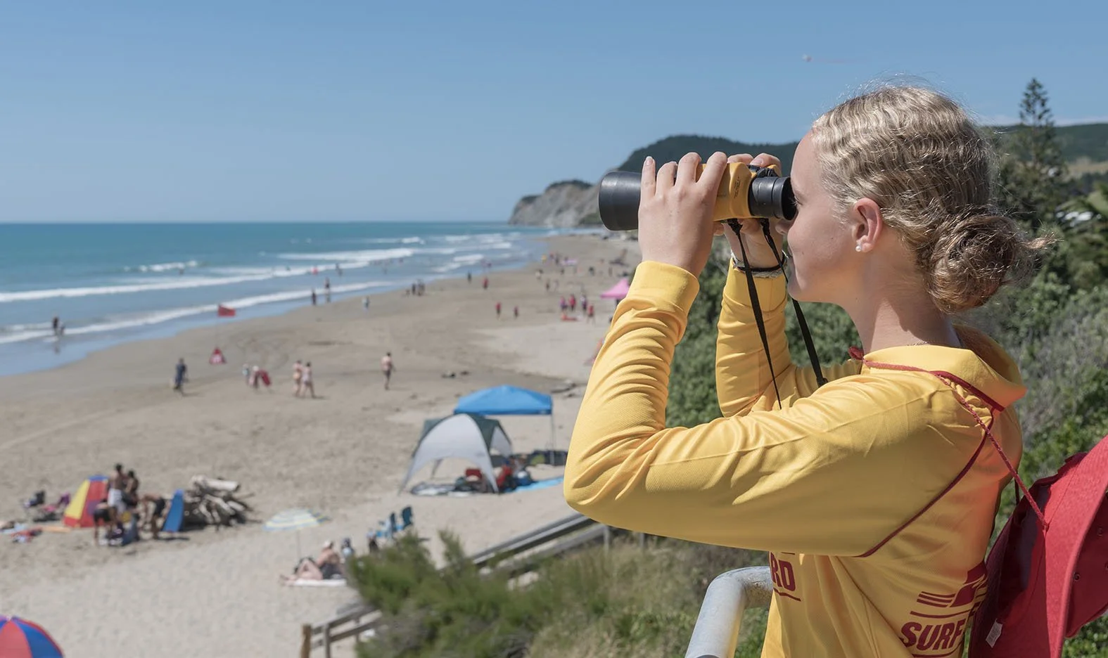 Woman in yellow jacket using binoculars on a scenic overlook at the beach with people, umbrellas, and waves in the background.