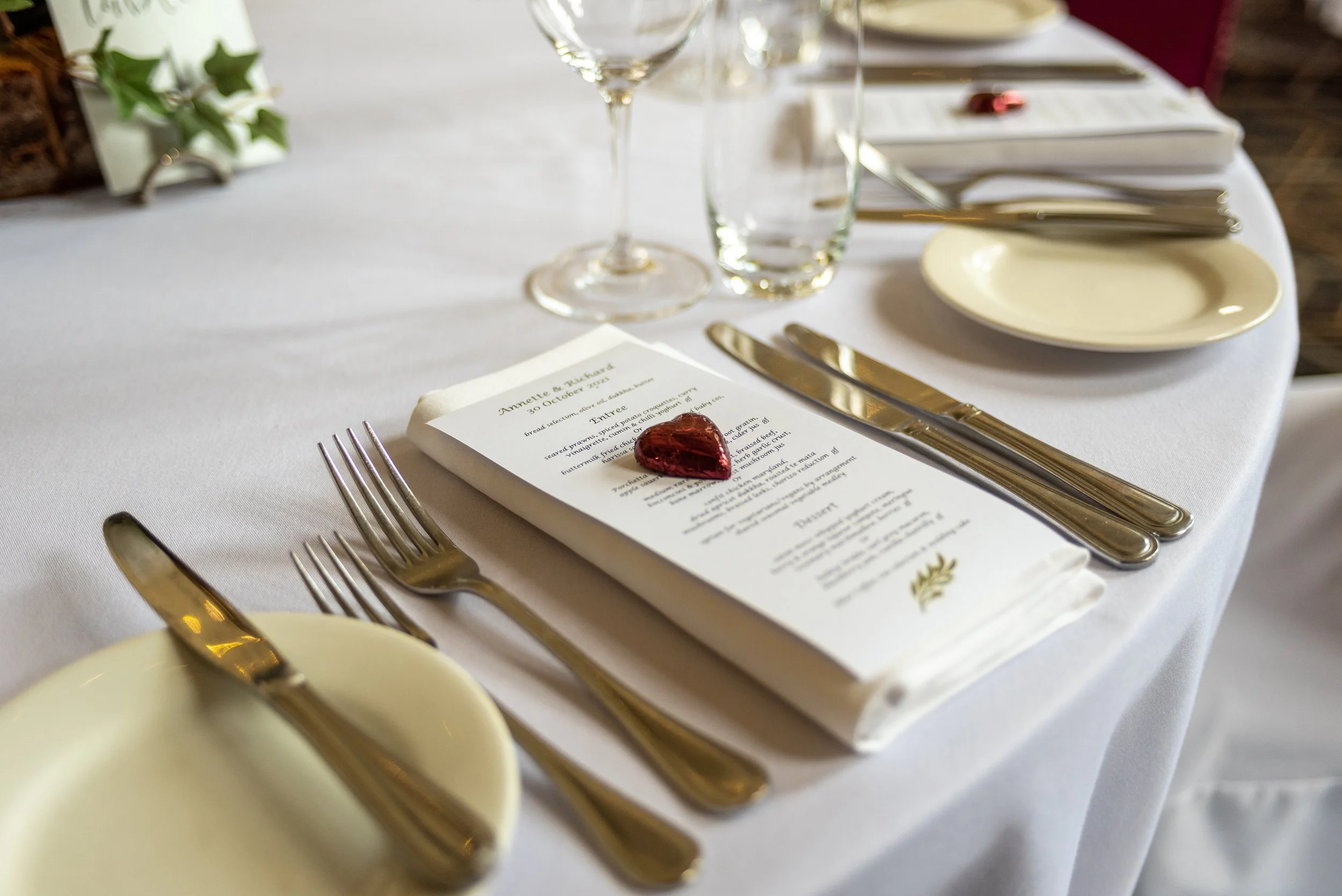 A wedding reception table setup with a menu card, gold utensils, wine glass, water glass, small plates, and a decorative heart-shaped item, all on a white tablecloth.