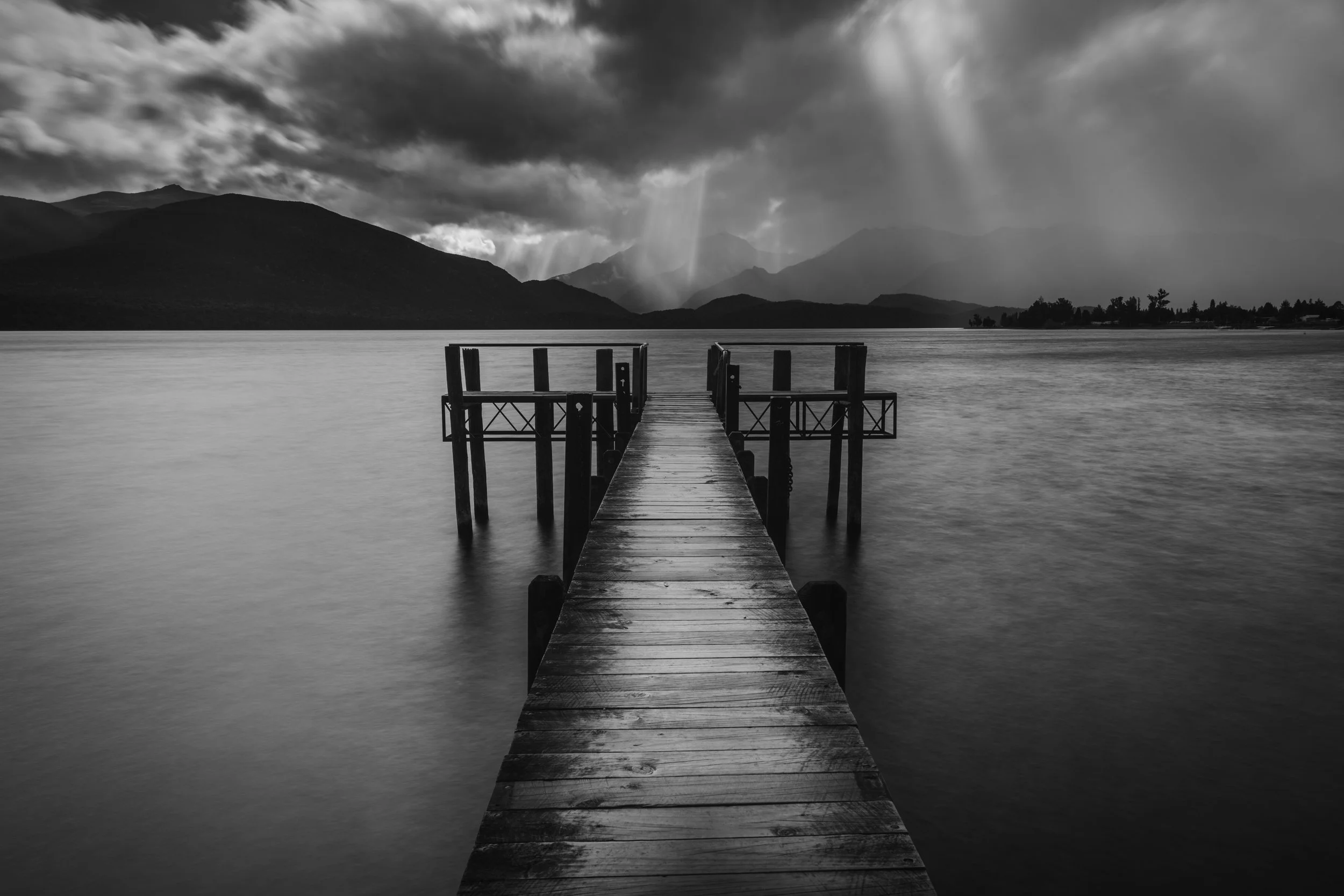A black and white photo of a wooden dock extending into a calm body of water, with mountains and stormy clouds in the background.