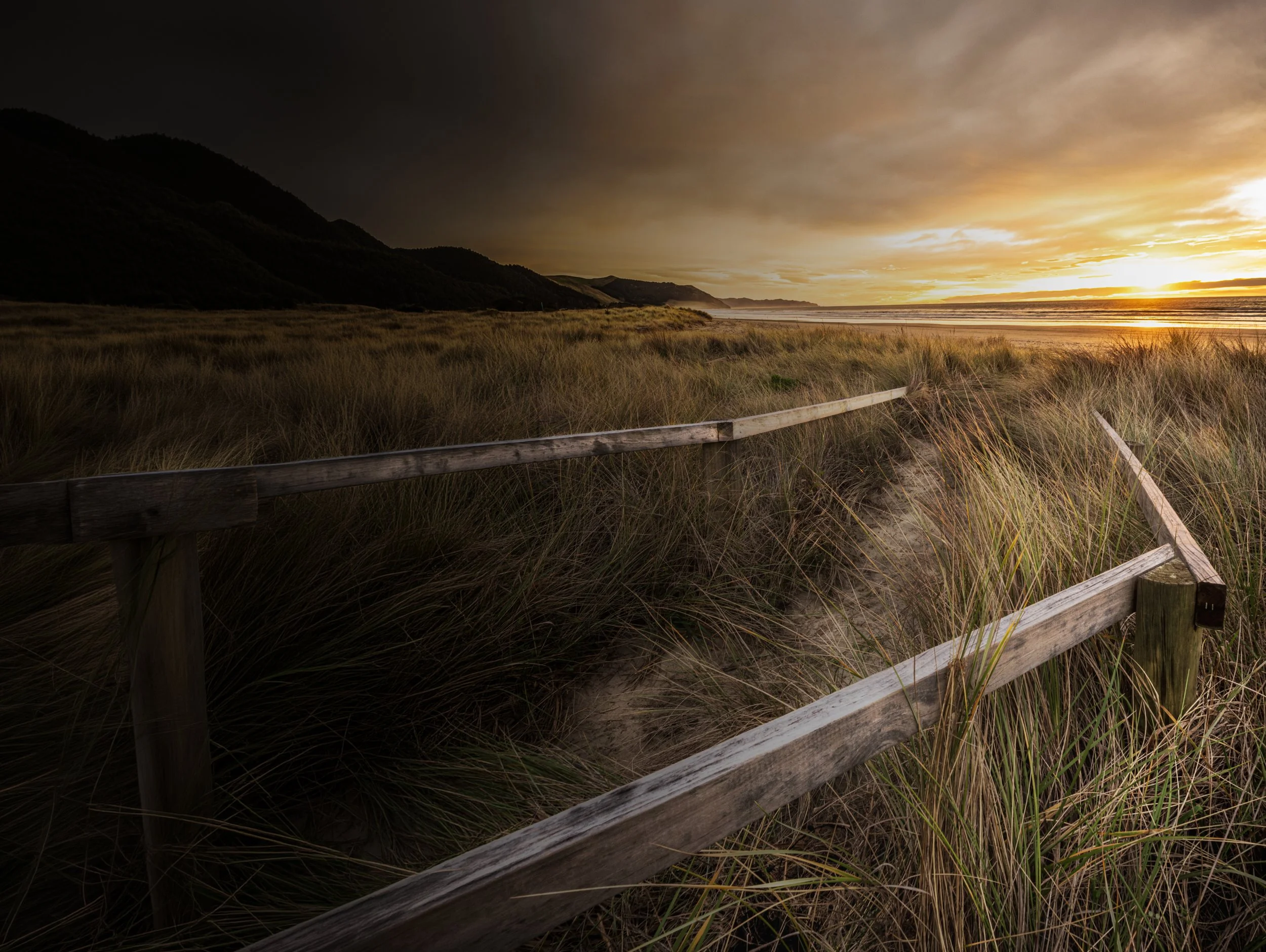 A beach landscape with dunes and tall grasses, a wooden fence, mountains in the background, and a sunset or sunrise with clouds and golden light