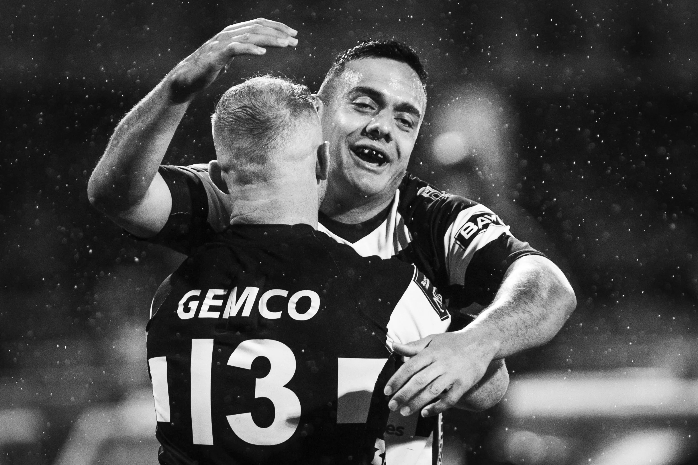 Two rugby players are hugging each other on the field, celebrating after a match in the rain. One player has a visible number 13 on his jersey and the word GEMCO. They look happy and are smiling.