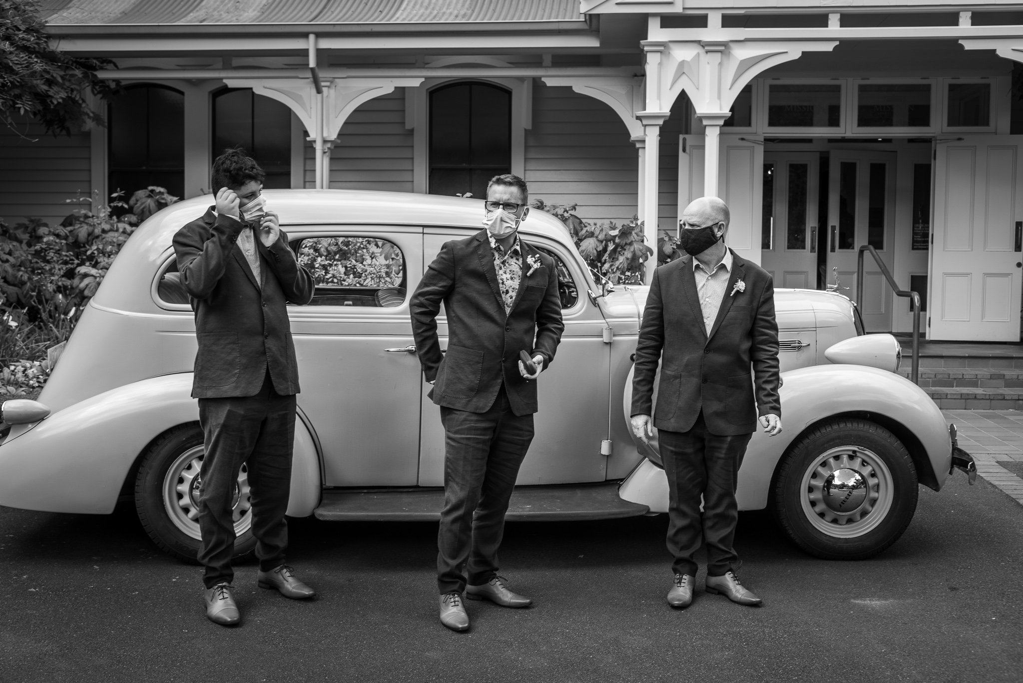 Three men in suits and masks standing in front of a classic car outside a house, with the man on the left adjusting his mask, and the man in the center holding a phone.