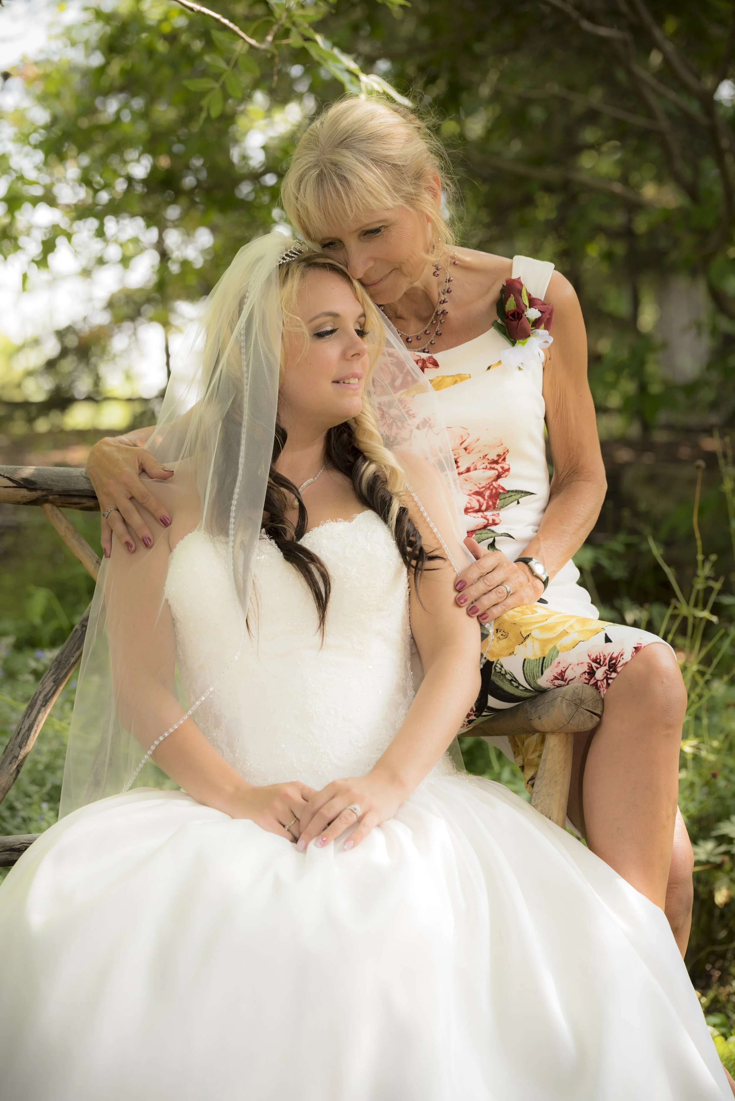 A bride in a white wedding gown and veil sitting outdoors with an older woman, possibly her mother, gently leaning her forehead against the bride's head. The older woman wears a white dress with a floral pattern, a necklace, and a corsage of red and 