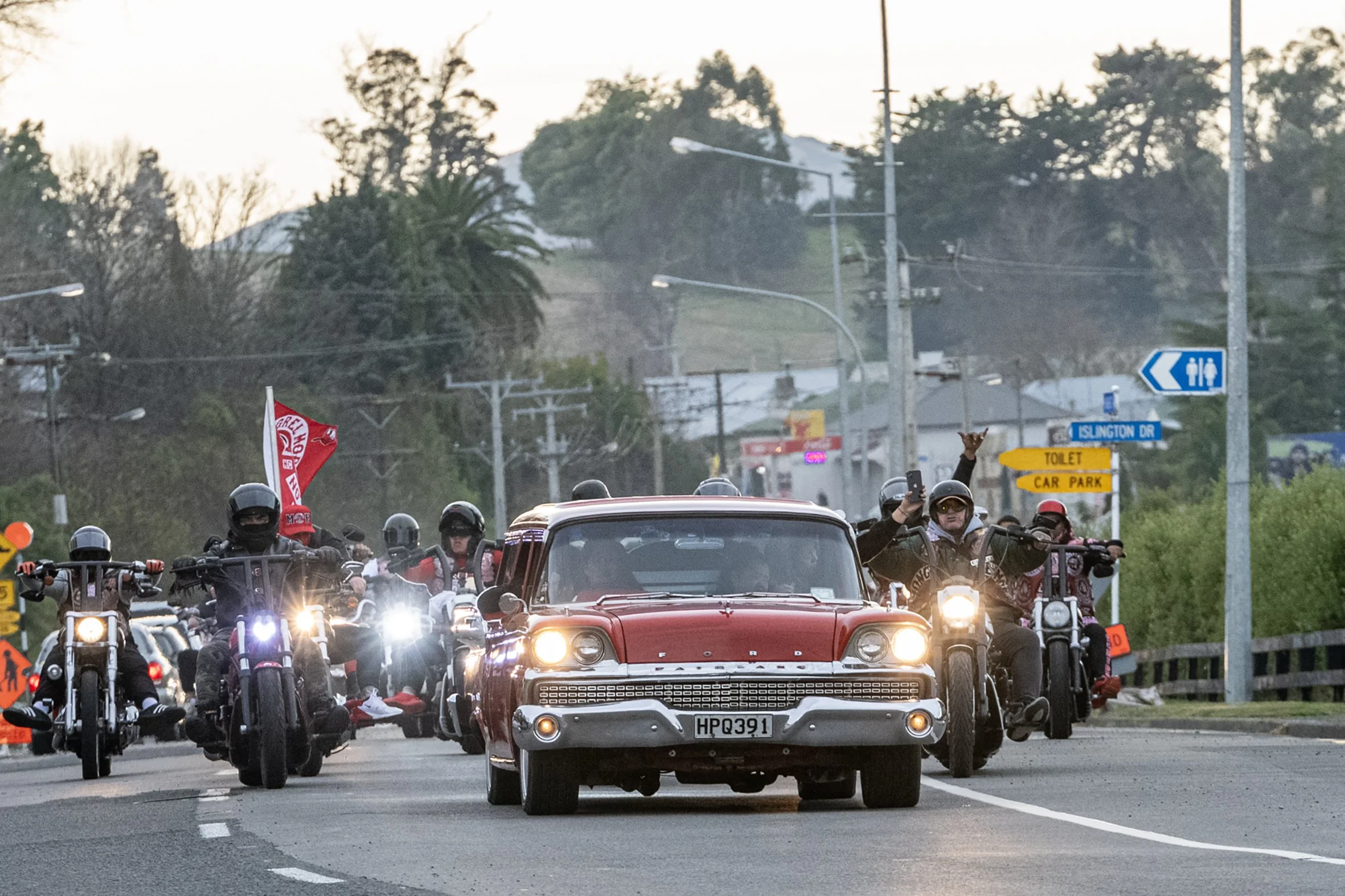 A group of motorcyclists riding together in a convoy, with a red vintage car in the front, traveling on a road with street signs and utility poles visible in the background during daylight.