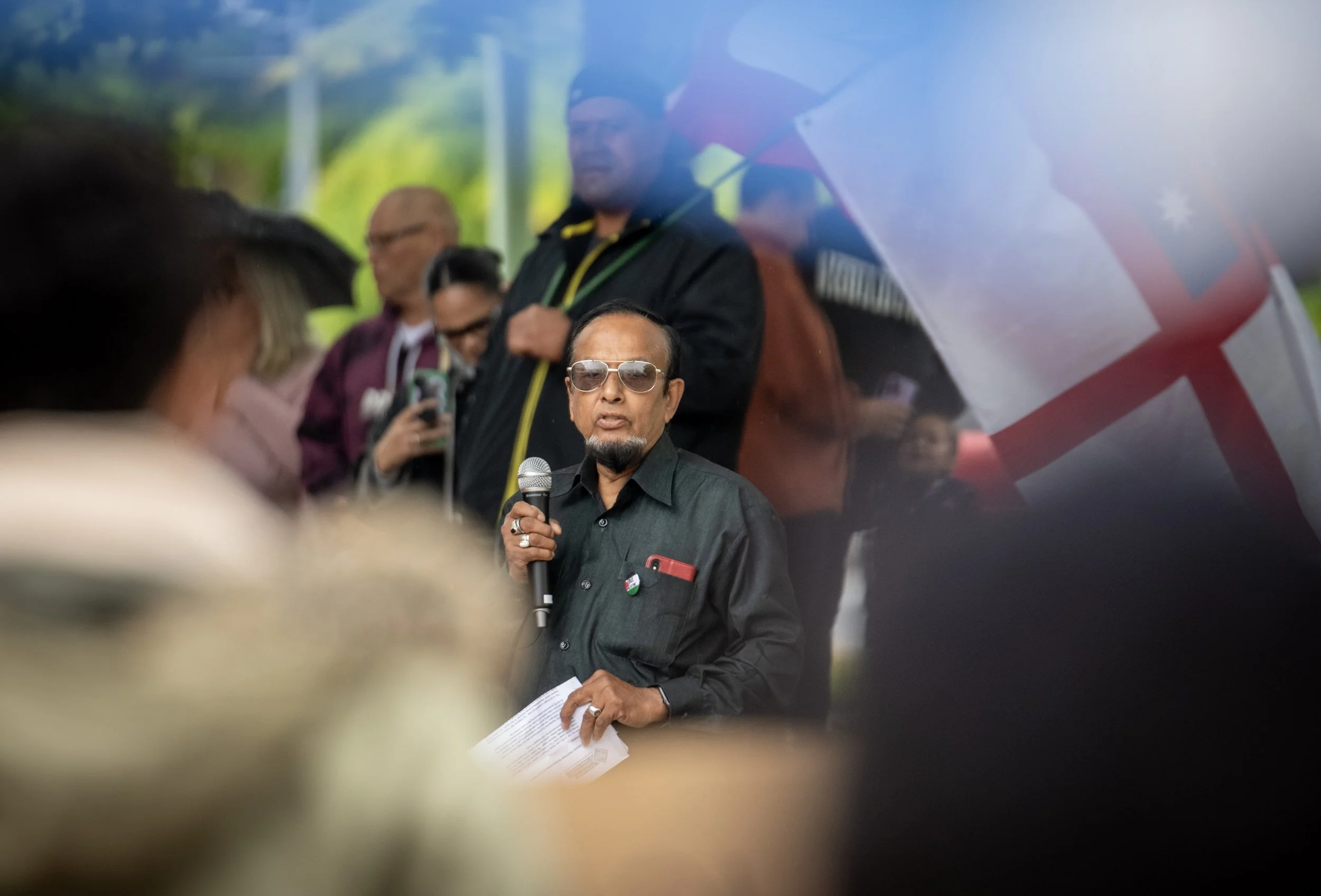 A man in dark clothing and sunglasses speaks into a microphone at a public event, with a crowd and a large flag in the background.