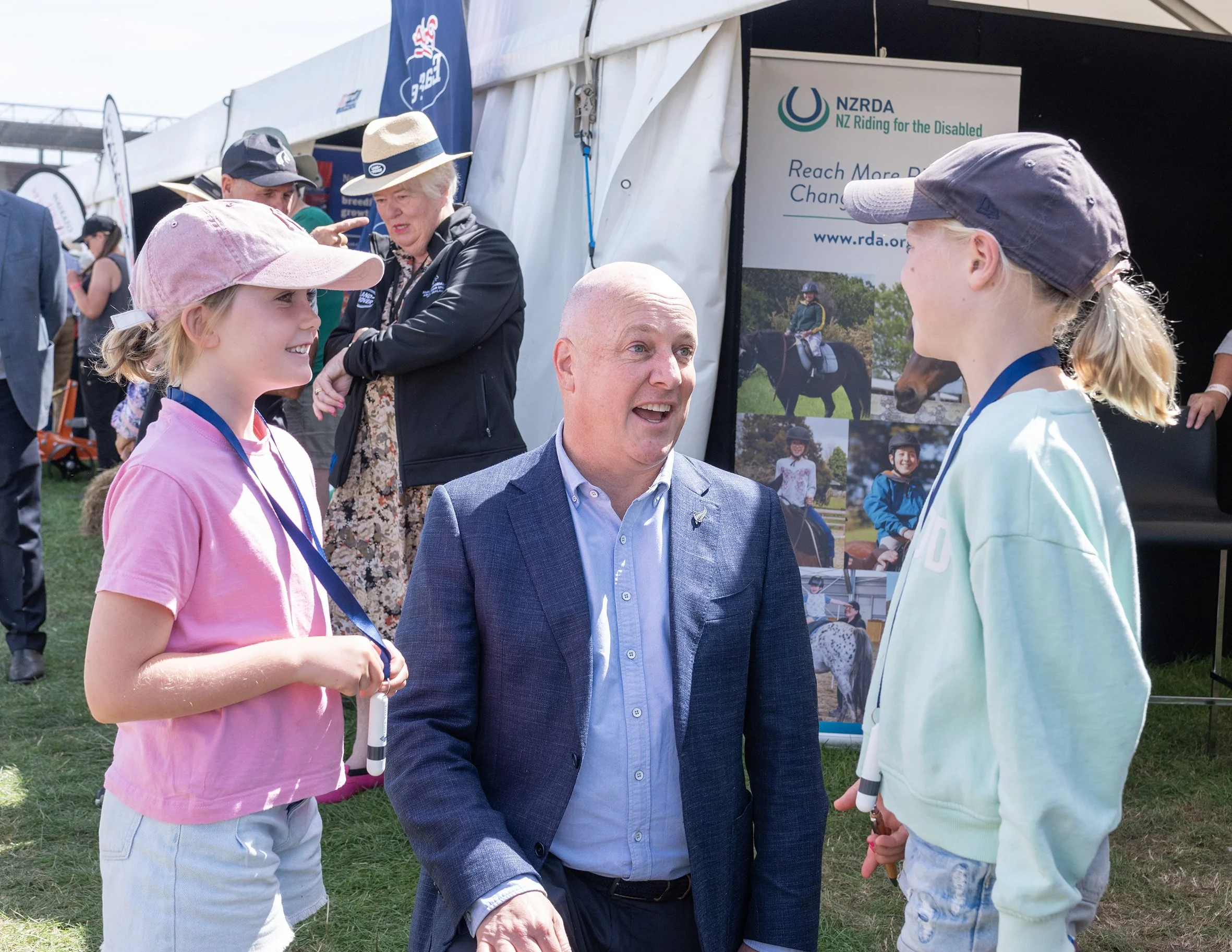 A group of four people, two young girls, an older woman, and a man, are talking outdoors at an event with tents and banners promoting NZ Riding for the Disabled. The girls are wearing caps, and the woman is wearing a broad-brimmed hat. They are engag