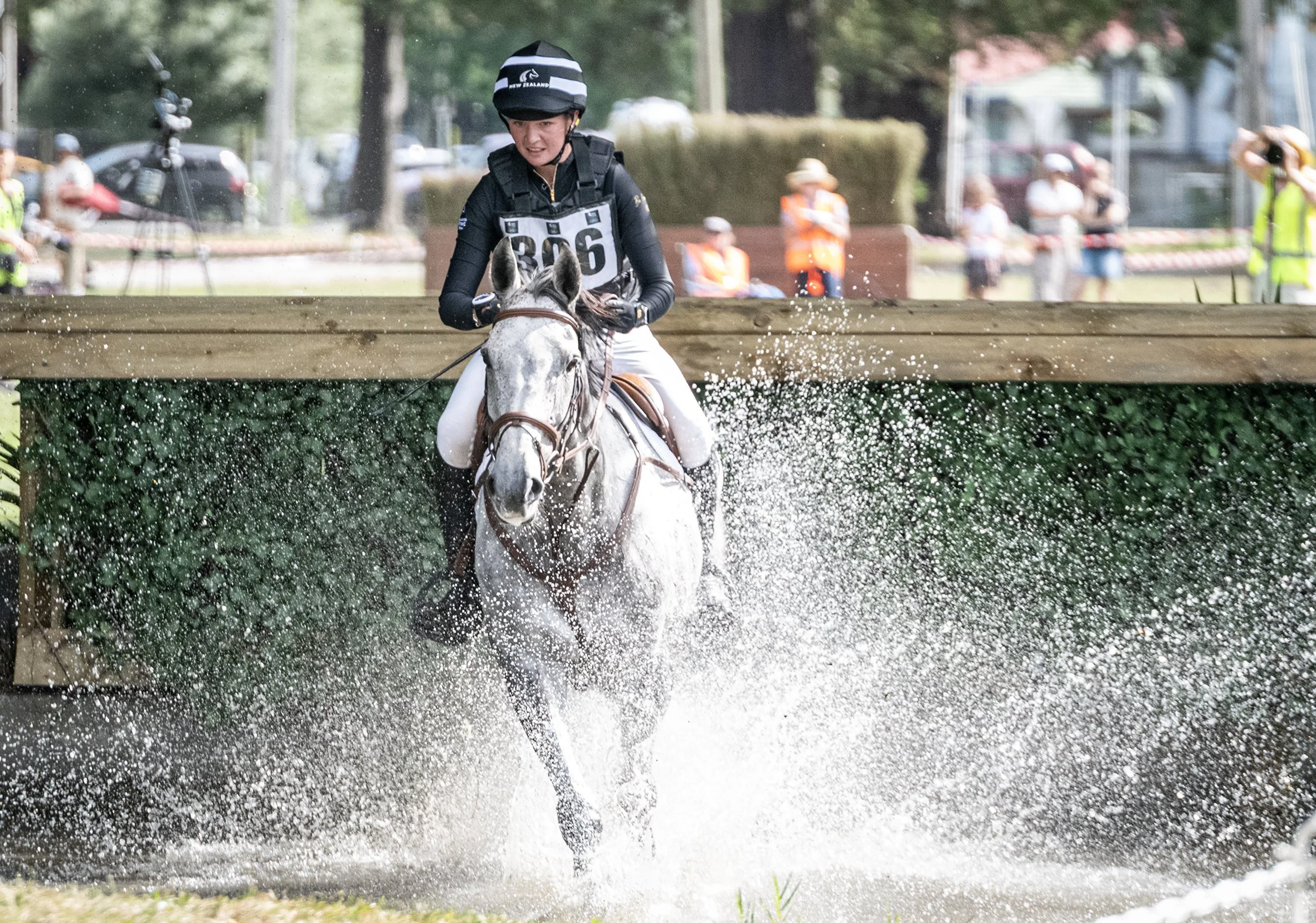 A person riding a horse over a water obstacle in an equestrian event, with spectators and officials watching in the background.