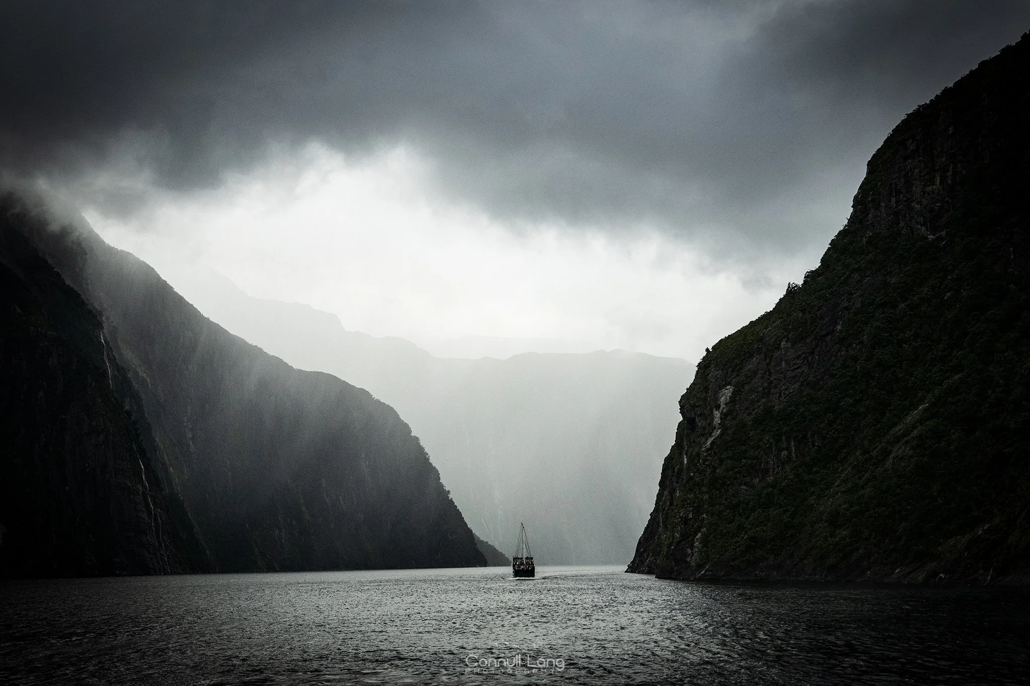 A boat sailing through a narrow, dark waterway between steep, forested mountains on a cloudy day