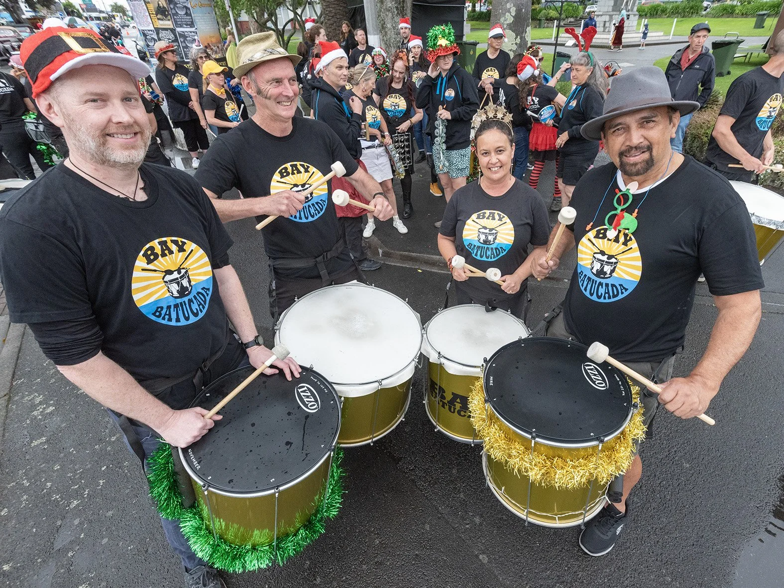 Group of four percussionists with drums decorated with green and gold tinsel, smiling during a festive parade, wearing black T-shirts with a drum logo and the words 'Bay Batu Cada,' and some wearing festive holiday hats.
