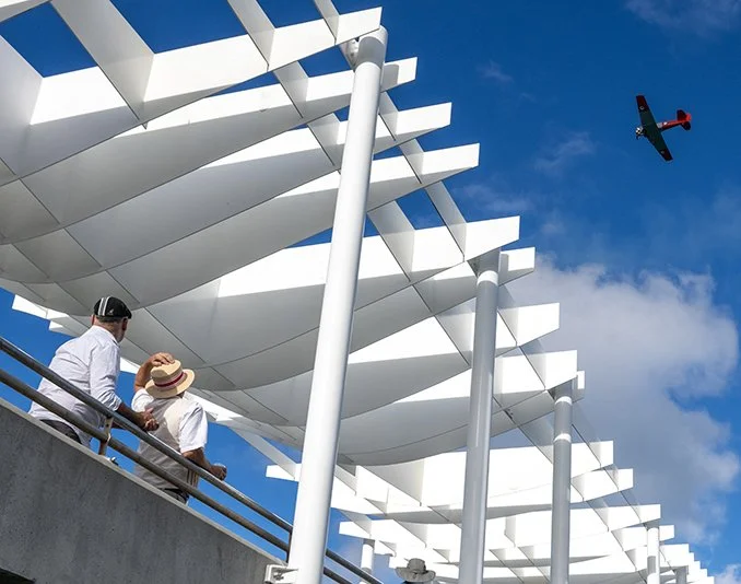 Two people walk on a bridge with a modern white architectural structure above, under a blue sky with some clouds, and a small airplane flying nearby.