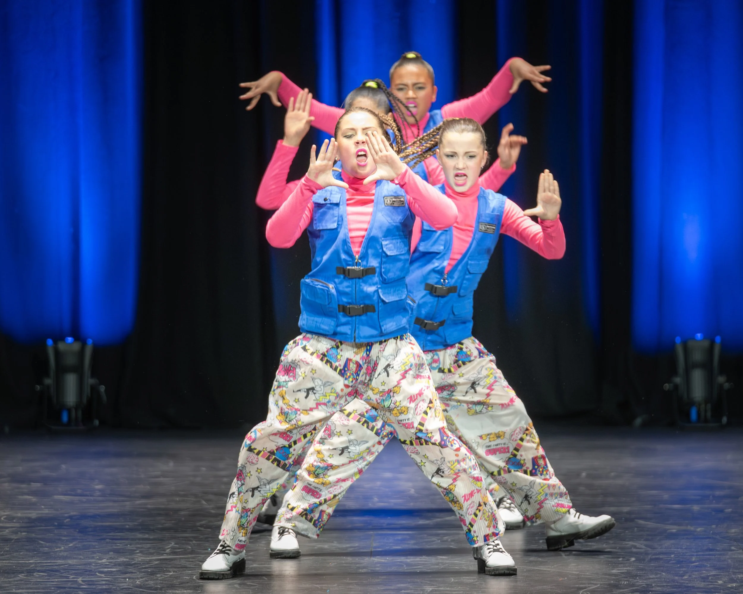 Four young female dancers in colorful outfits performing on stage with a dark background and blue curtains.